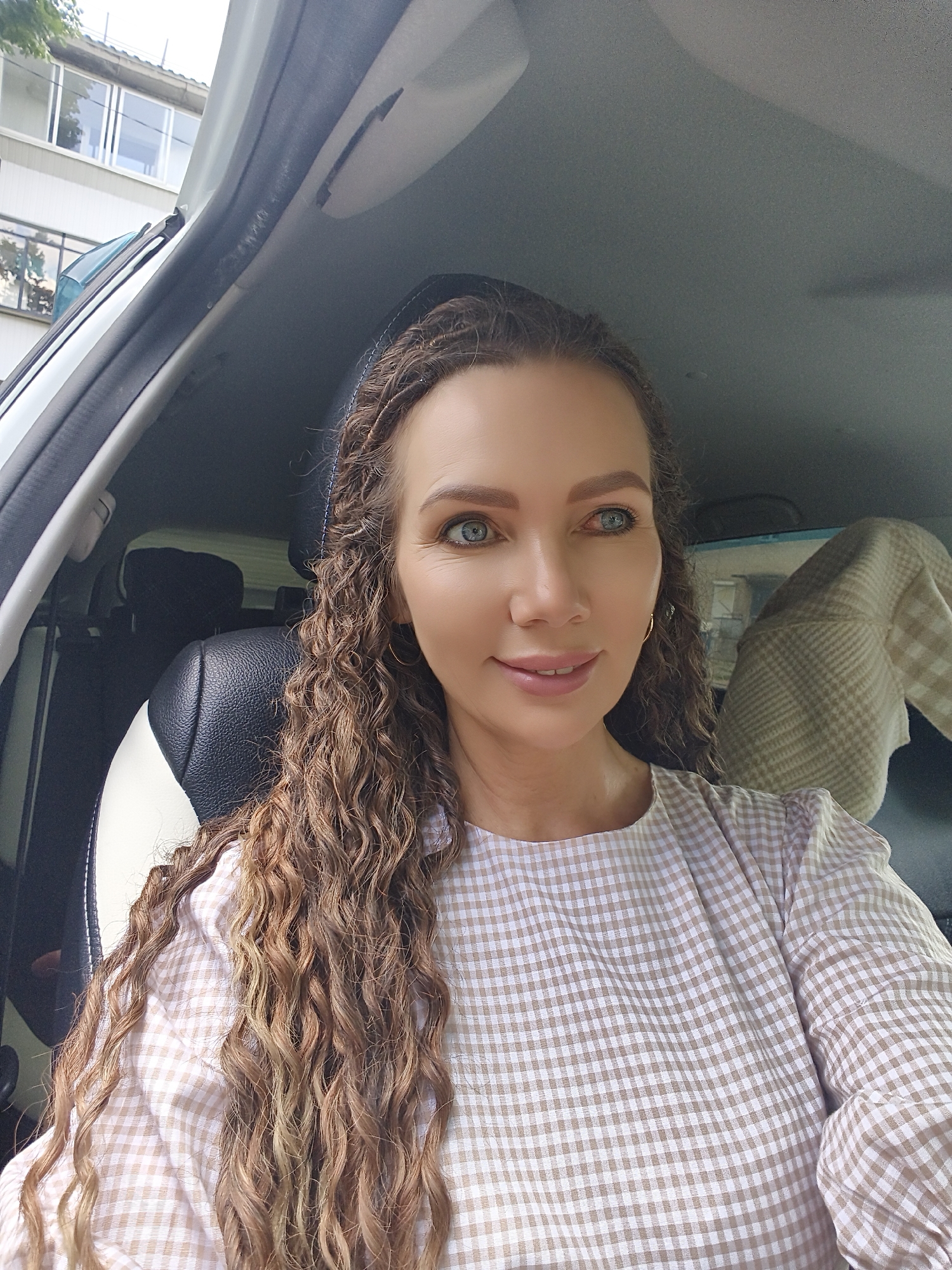 A woman with long, curly brown hair and blue eyes is sitting in the driver's seat of a car. She is wearing a beige and white checkered shirt. The interior of the car is visible, including the car seat and part of the dashboard. Outside the car, a building with windows can be seen. The scene appears to be outdoors, possibly in a residential area.