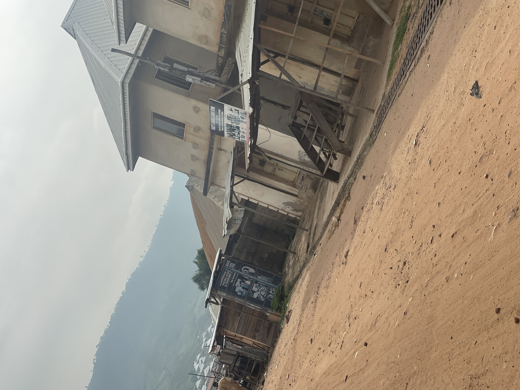 The image captures a street scene in Lokoja, Nigeria, characterized by a dirt road and buildings with corrugated metal roofs. The structures appear to be a mix of shops and residential spaces, some of which are under construction or in a state of disrepair, with visible wooden scaffolding. Signs are present on some of the buildings, including one for "VICKY'S LIQUOR & MORE STORE" and another for a phone repair and charging center. In the background, a lush green hillside is visible, shrouded in mist, suggesting a slightly elevated or mountainous terrain surrounding the city. The sky is overcast, contributing to a muted and slightly hazy atmosphere. There are no people clearly visible in action, but a person in light-colored clothing is partially seen walking on the left side of the image. The overall scene depicts a typical, somewhat rustic urban environment in Nigeria.