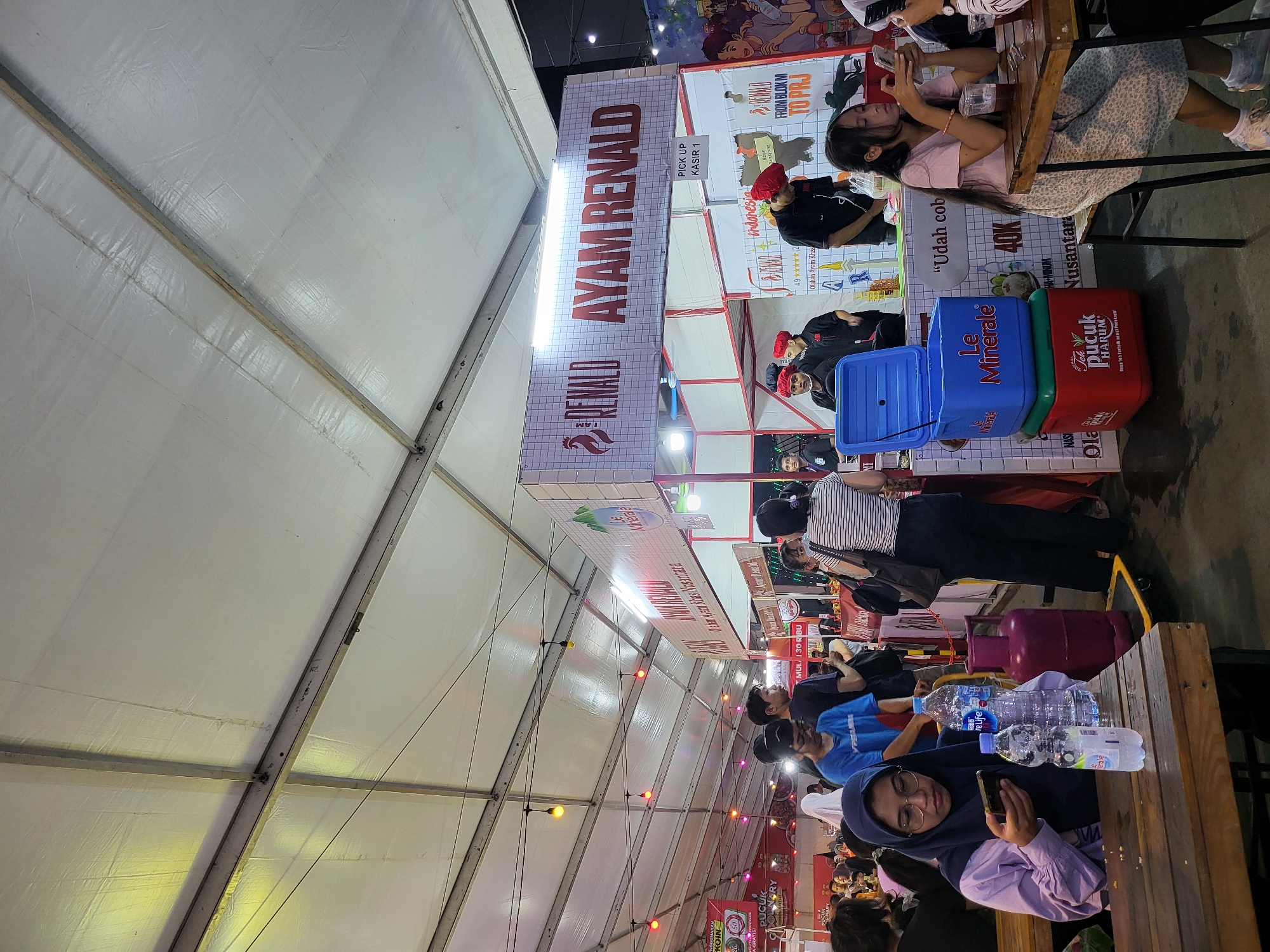 The image depicts a bustling food stall at what appears to be an outdoor market or festival in North Jakarta, Indonesia, under a large white tent structure. The stall is branded "AYAM RENALD" and seems to specialize in fried chicken. Several people are visible, including customers and staff.
In the foreground, a young woman wearing a hijab and glasses is seated at a wooden table, looking at her mobile phone. Two bottles of water and a large pink gas cylinder are also on the table. To her right, another young woman in a pink top is seated, also looking at her phone, with a glass of iced tea on the table.
Behind them, the "AYAM RENALD" stall is set up, with staff members wearing red caps and black shirts preparing food. A large blue cooler box labeled "Le Minerale" and a red cooler box labeled "Teh Pucuk Harum" are stacked on the counter. Banners with Indonesian text are visible, including one that says "AYAM RENALD" and another with "PICK UP KASIR 1".
The environment suggests a vibrant evening event, with string lights illuminating the tent ceiling and various food and drink vendors in the background. The overall atmosphere is lively and communal, typical of a local market setting.