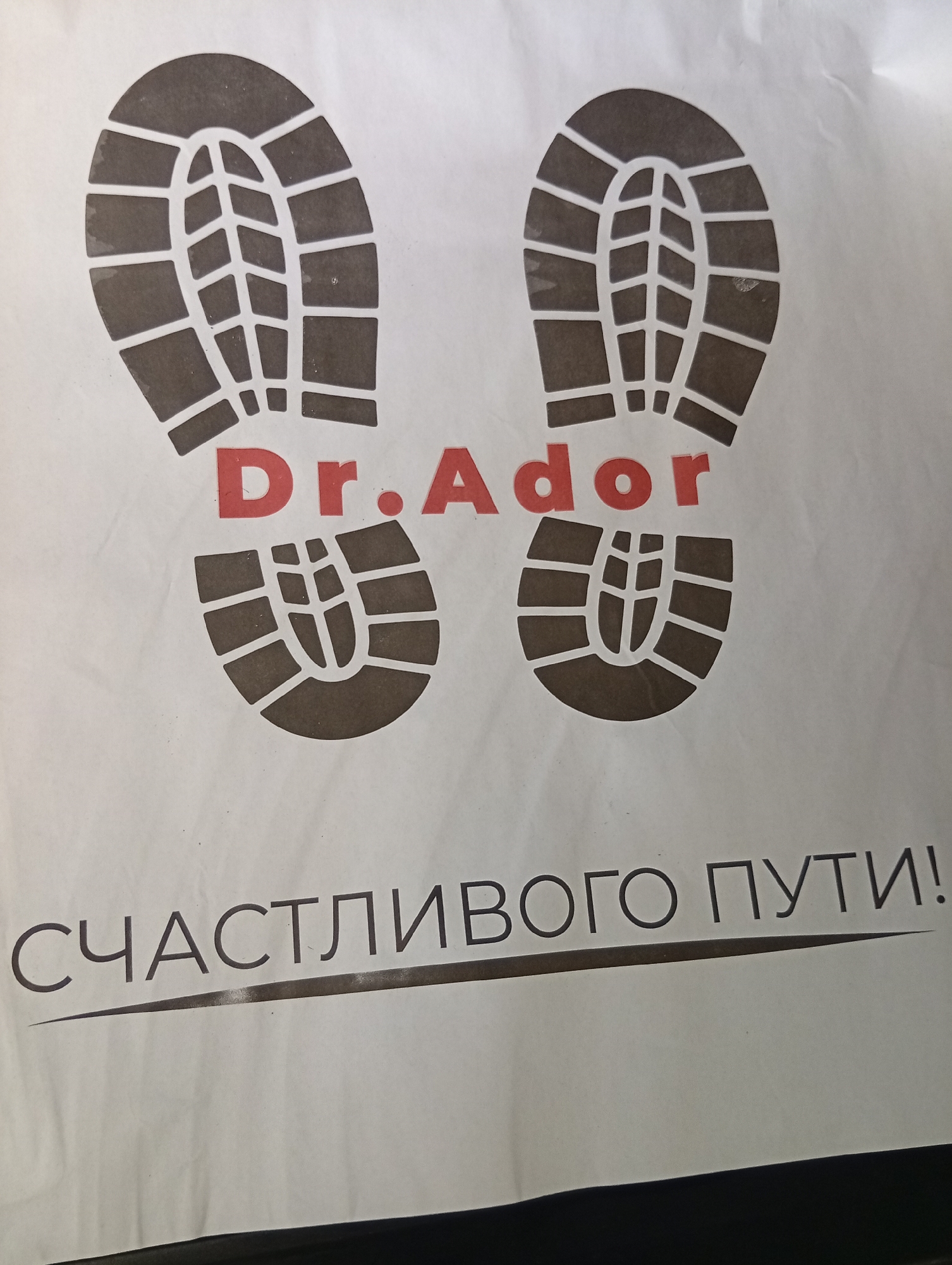 The image shows a white paper with two boot prints and text in Russian. The text reads "Dr.Ador" in red, and below it, "СЧАСТЛИВОГО ПУТИ!" (Happy journey!). The boot prints are dark brown. The text and boot prints are printed on a white background. The lighting appears to be natural, and the image is taken from a slightly upward angle. There is no visible indication of the specific location of the image in Tashkent, Uzbekistan, or any people or specific actions taking place.