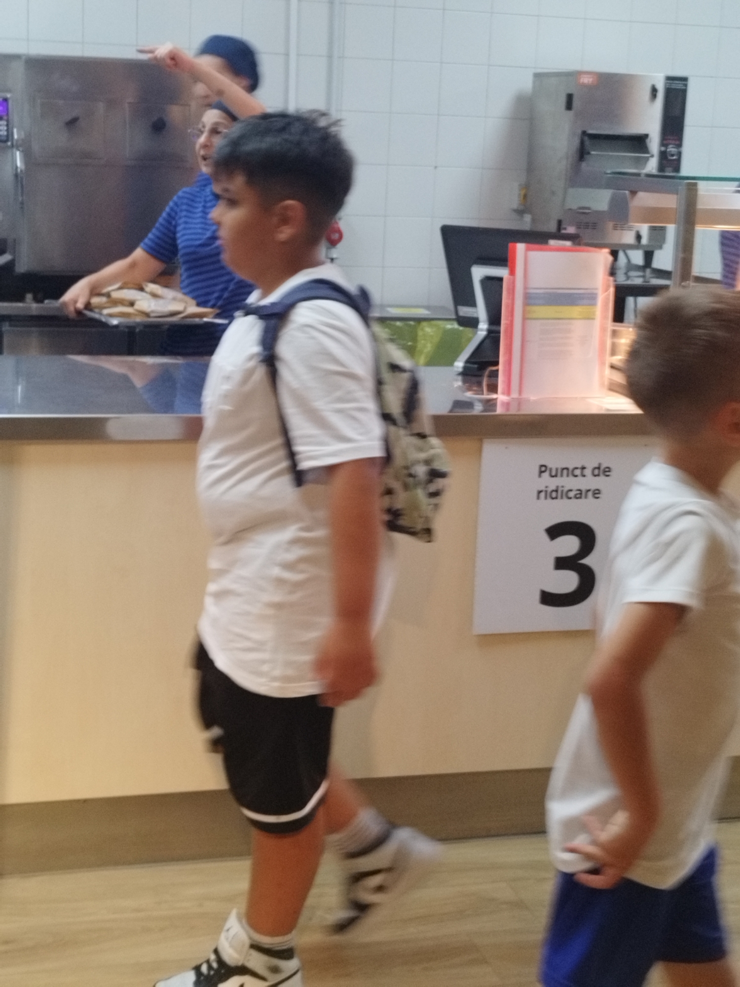 This image captures a scene inside a food service establishment, likely a cafeteria or canteen, with a distinct Romanian context suggested by the signage. In the foreground, a young boy wearing a white t-shirt, black shorts, and white sneakers with black accents is walking. He has a backpack with a camouflage pattern slung over his right shoulder. To his right, another child, partially visible from the side, is wearing a white polo shirt and blue shorts. In the background, a woman wearing a blue striped shirt and a dark blue hairnet is seen serving food from a tray. Another person, wearing a blue cap, is pointing upwards. The environment is characterized by white tiled walls, stainless steel serving counters, and a visible sign with Romanian text reading "Punct de ridicare 3," which translates to "Pick-up point 3." The overall lighting suggests an indoor setting with artificial illumination.