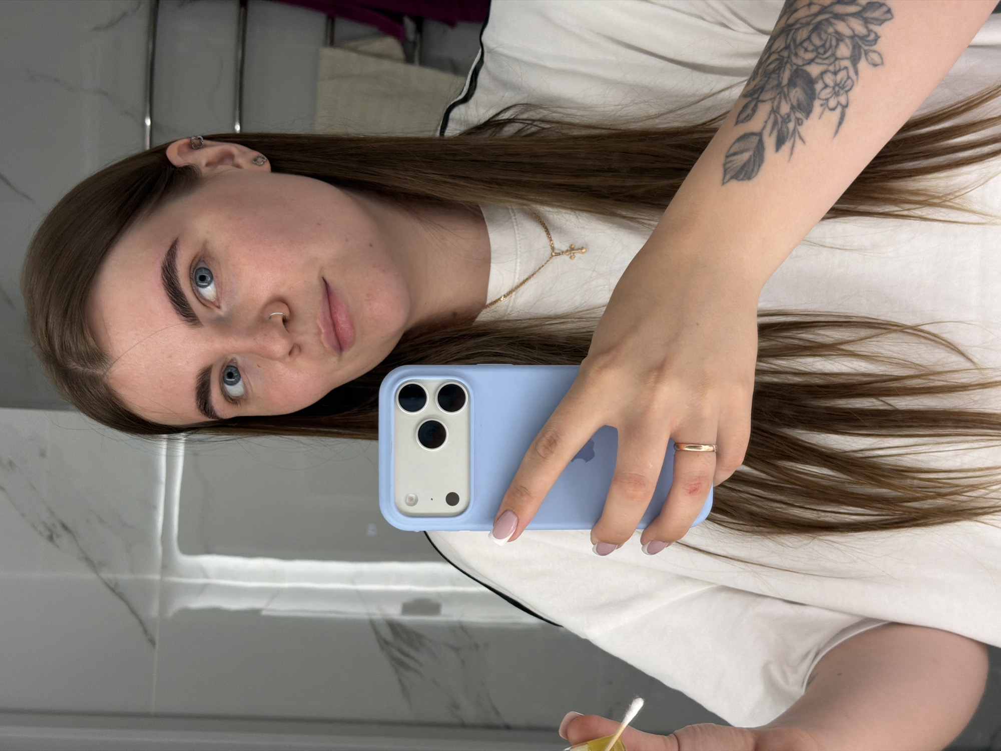 A woman with light skin, brown hair, and blue eyes takes a selfie in what appears to be a bathroom. She is wearing a white t-shirt and a gold necklace with a cross pendant. Her left arm is visible, showing a floral tattoo. She is holding a blue smartphone in her right hand, displaying manicured fingernails. A gold ring is on her ring finger. In her other hand, she holds a cotton swab. The background shows tiled walls, a mirror, and some hanging items. There is no indication or evidence that this image was taken in the city of Chaikovsky, Russia.
