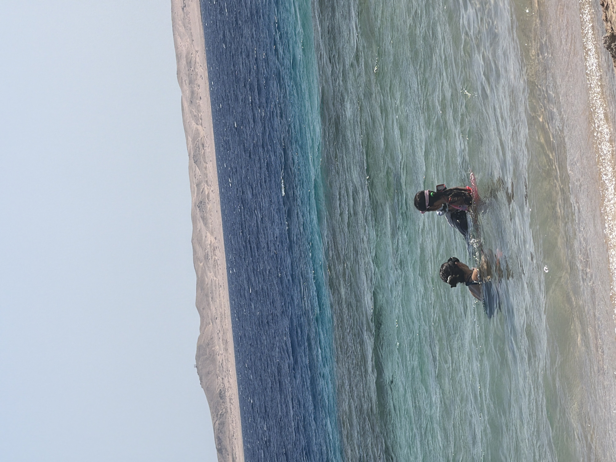 In Sharm El Sheikh, Egypt, two individuals are present in the water near the shore. The person on the left is wearing a dark swimsuit and has their hair tied back. The person on the right is equipped with snorkeling gear, including a mask and a snorkel, and is wearing a dark suit with a pink flotation device visible. The sea exhibits distinct color variations, transitioning from a lighter turquoise near the shore to a deeper blue further out. In the background, a barren, sandy mountain range is visible under a clear, light blue sky. The water is slightly choppy, with small waves reaching the sandy beach in the foreground.