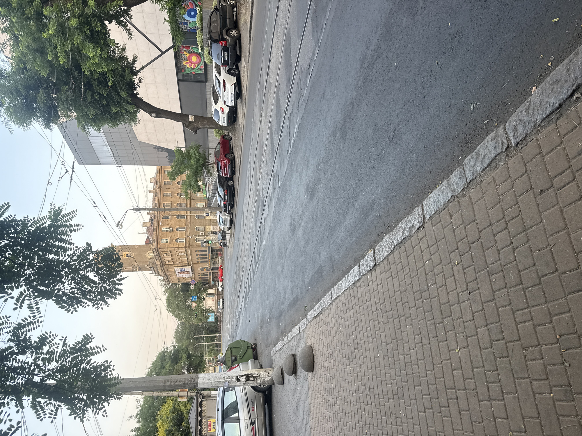 The image captures a street scene in Odesa, Ukraine, likely during daytime with overcast skies. The perspective is from a sidewalk, looking across a street with tram tracks. On the left side of the street, a silver car is parked next to a tall, grey utility pole. A large green dumpster is situated behind the utility pole. The sidewalk is paved with rectangular brown bricks, bordered by a stone curb. Several round, grey concrete bollards are placed along the edge of the sidewalk.
Across the street, a row of parked cars, including a black SUV, a white sedan, a red sedan, and several other vehicles, are visible. In the background, there are buildings, one of which is a multi-story structure with a clock tower and tan-colored facade. Another building on the right appears more modern with a grey, glass facade and a colorful mural on its lower section. Trees with green foliage are scattered throughout the scene, some overhanging the street and sidewalk. The street itself is asphalt, appearing somewhat worn with visible tram tracks. The overall impression is of an urban environment with a mix of older and newer architecture.