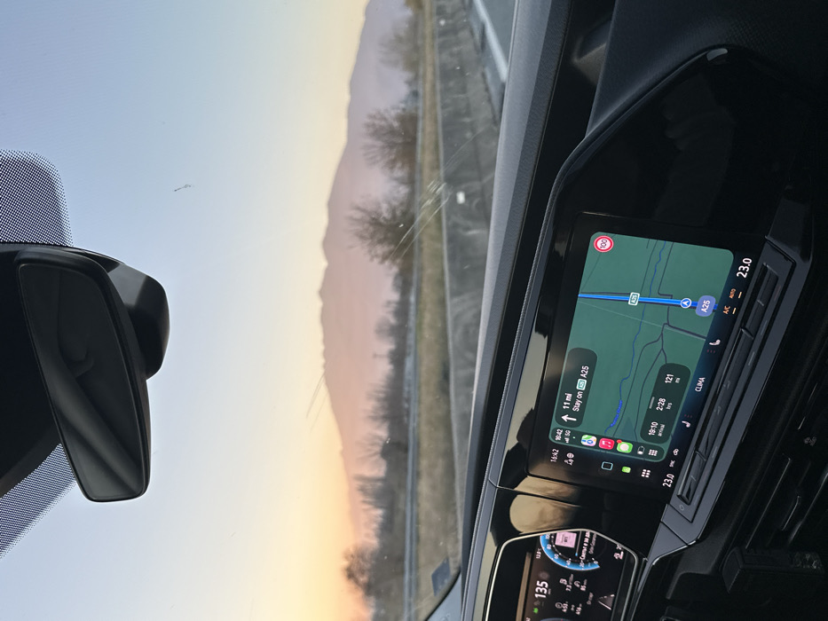 The image presents a forward-facing view from inside a moving vehicle on a highway, identified as the A25 in the vicinity of Sidirokastro, Greece. The exterior scene depicts a clear sky transitioning from pale blue overhead to warm orange and pink tones near the horizon, indicative of either sunset or sunrise. A distant, hazy mountain range forms a prominent silhouette against this colorful sky. The highway itself features multiple lanes separated by a concrete barrier, with sparse, dry vegetation lining the roadside.
Inside the vehicle, a modern dashboard is visible. The digital instrument cluster on the left displays a speed of 135 (presumably km/h). A central infotainment screen shows a navigation map with the vehicle positioned on the A25 highway, displaying a current time of 16:42, a remaining distance of 11 miles, an estimated arrival time of 18:10, and approximately 2 hours and 121 minutes of travel time. A 100 km/h speed limit sign is visible on the map. Climate control settings indicate 23.0 degrees Celsius. A rearview mirror assembly is affixed to the top of the windshield, which exhibits minor streaks and a small impact point or crack in the lower-central area. No individuals are visible within the frame.