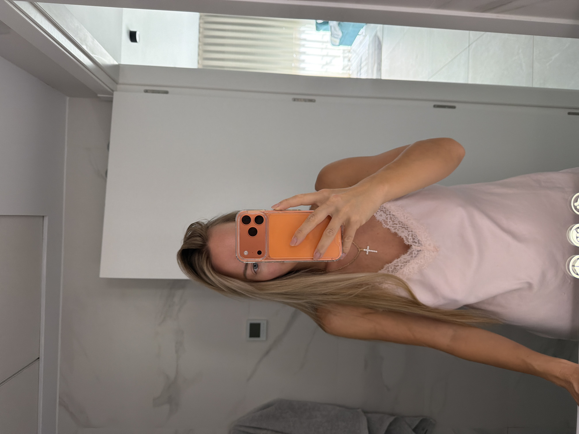 A woman with long blonde hair takes a mirror selfie in a bathroom. She is wearing a light pink slip with lace trim and a cross necklace. She is holding an orange phone in her right hand, obscuring part of her face. The bathroom has marble walls and a white door. In the reflection of the mirror, a room with light curtains and a blue sofa can be seen. The image appears to have been taken in Finestrat, Spain.