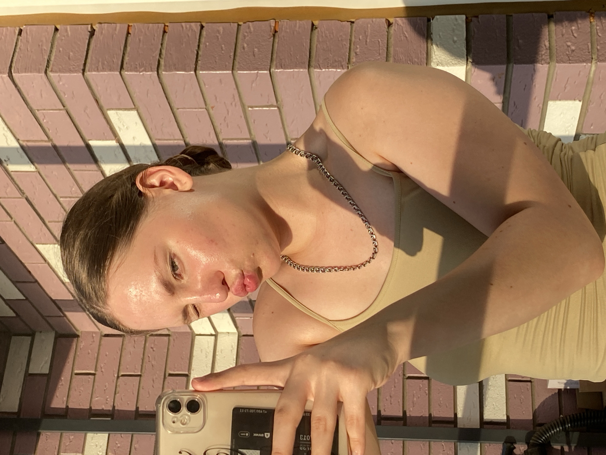 A young woman is taking a selfie outdoors, with a brick wall in the background. The wall is made of pink and white bricks and has a shadow cast across it. The woman is wearing a beige tank top and a silver chain necklace. She is winking and puckering her lips, and her phone is visible in her hand. The lighting suggests it is either sunrise or sunset. This image was taken in Izhevsk, Russia.
