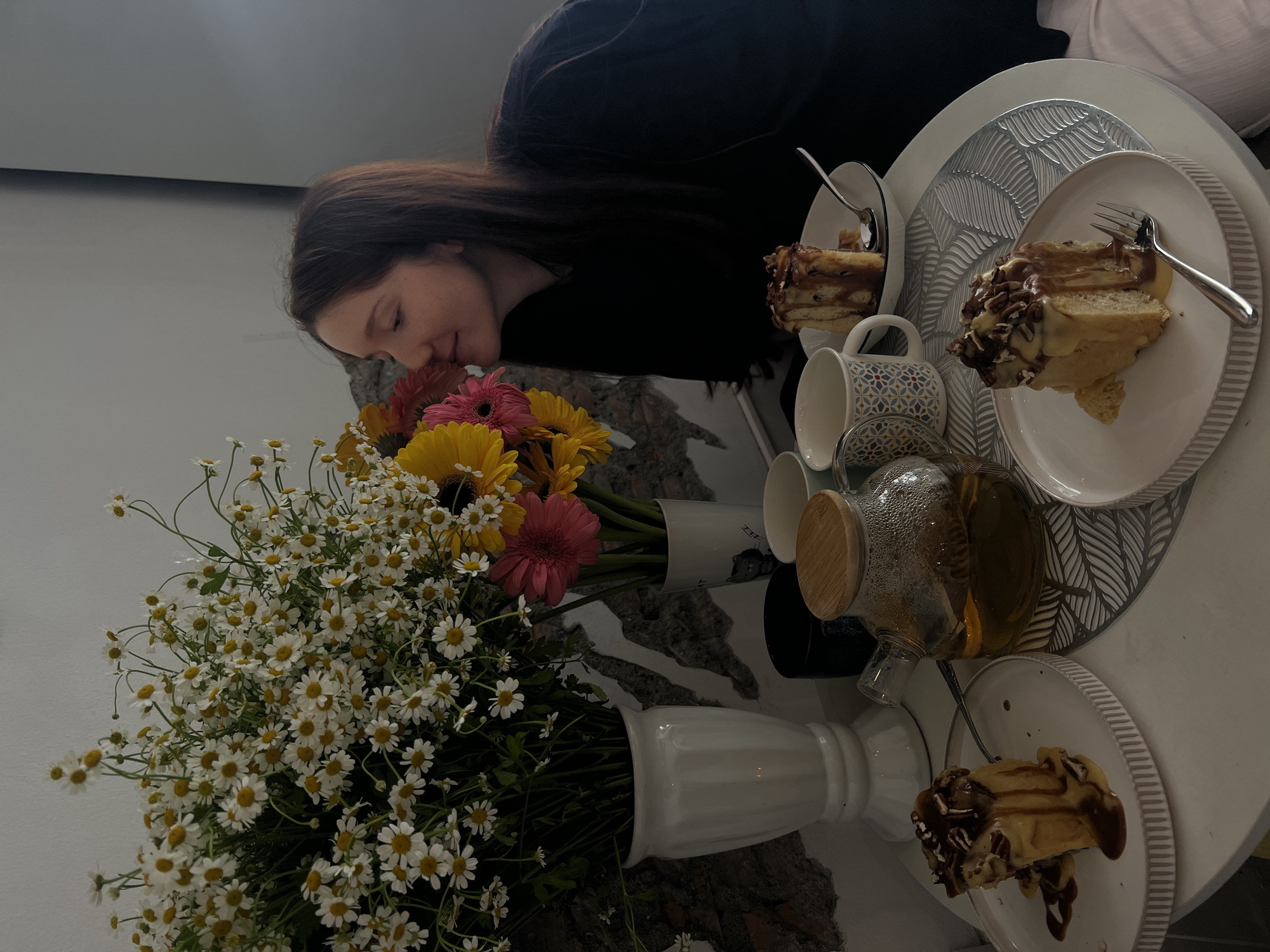 A Caucasian woman with long brown hair is bent over a bouquet of daisies and gerberas in vases on a round table. Next to the flowers are two mugs and a glass teapot containing liquid. Three plates of what appears to be sliced cake, each garnished with nuts and sauce, are arranged around the table, each with a fork or spoon. The table has a white top and a rough, textured base. The setting may be an interior space in Phuket City Municipality, Thailand, though specific landmarks are not visible.