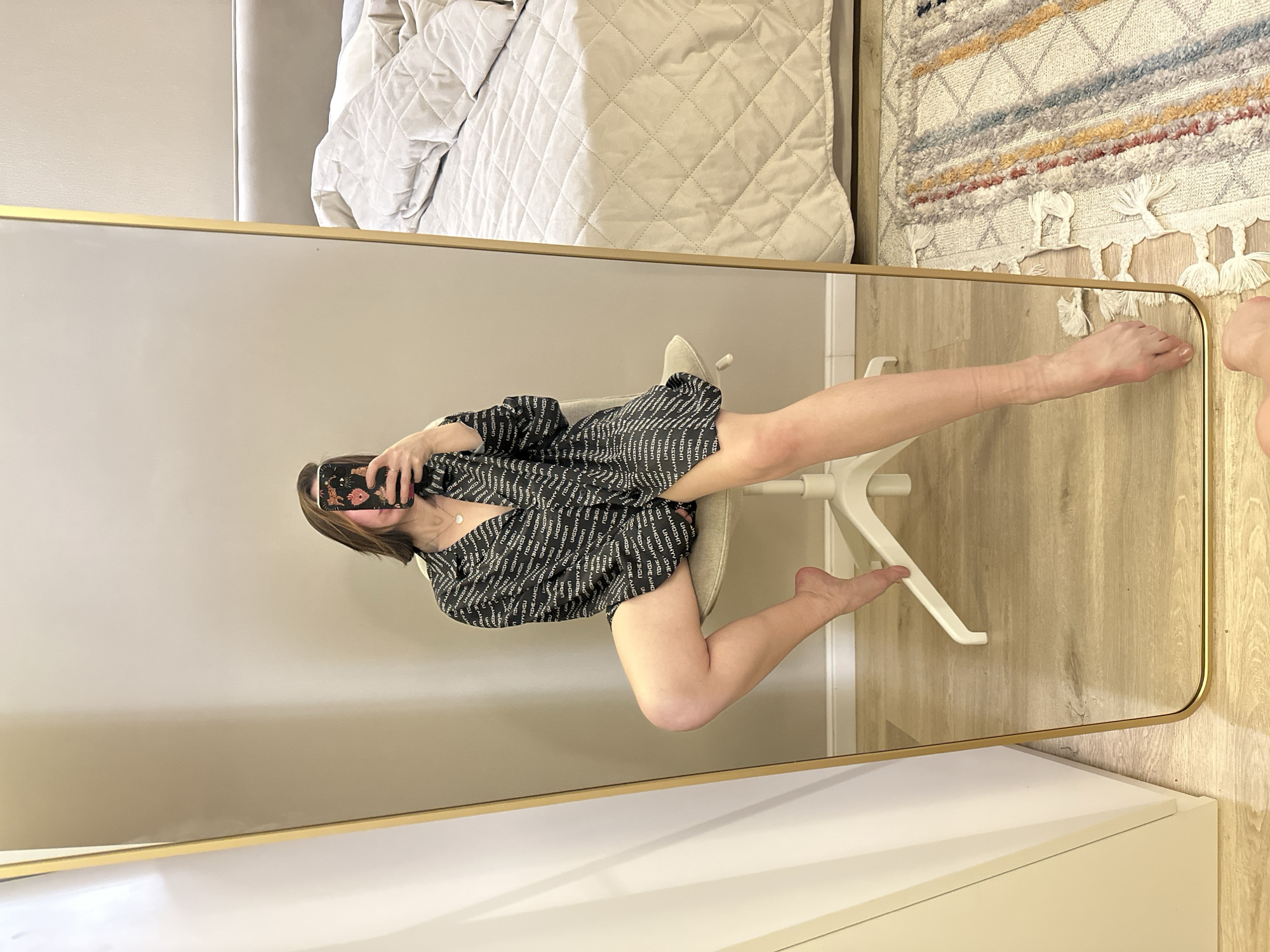 A young woman is reflected in a gold-framed mirror. She is sitting in a white office chair wearing a black and white patterned oversized shirt. She is holding a phone with a floral case in her hand, presumably taking a selfie. Her legs are positioned in an unusual, almost athletic pose, with one leg extended towards the top right corner of the mirror and the other bent under the chair. In the mirror's reflection, a corner of a bed with a neutral-colored quilted bedspread can be seen at the top, as well as a fringed rug. The floor appears to be light wood. The bottom of the mirror reflects a white cabinet. The location is not identifiable as being in Votkinsk, Russia, based on the visual information.
