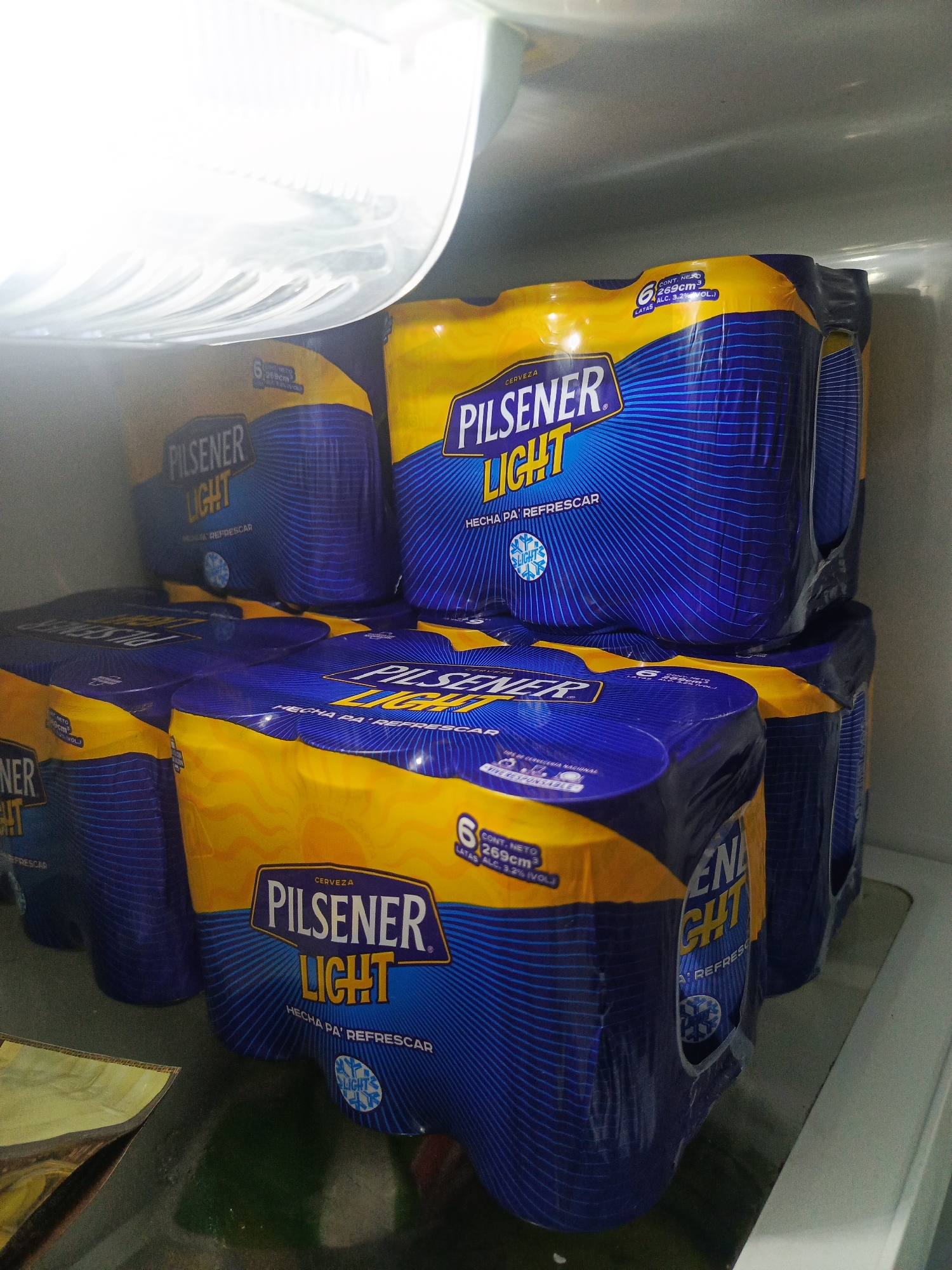 The image shows multiple six-packs of Pilsener Light beer inside a refrigerator. The beer packs are predominantly blue and yellow, with the Pilsener Light logo prominently displayed in white and blue lettering. Some packs also have text indicating "HECHA PA' REFRESCAR" (Made to refresh) and "CERVEZA" (Beer). The refrigerator interior is visible, including its white interior walls and a clear plastic shelf. The lighting suggests the refrigerator door is open. The scene is likely located within a home or a commercial establishment in Guayaquil, Ecuador, as Pilsener is a well-known Ecuadorian beer brand.