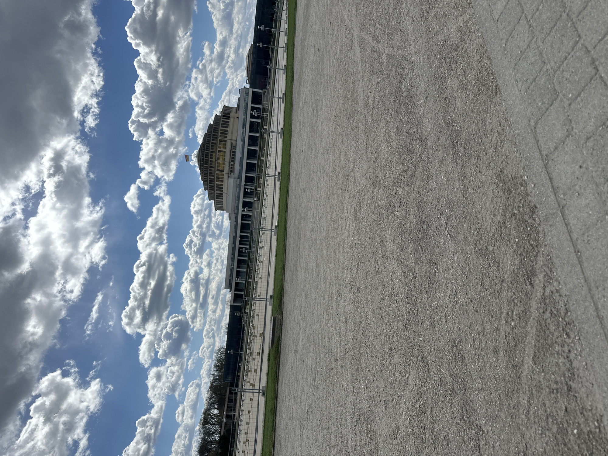 The image shows Centennial Hall in Wrocław, Poland, under a sky with cumulus clouds. The building, a multi-purpose structure, has a distinctive circular design with layered tiers. A flag is visible on the roof. The foreground consists of a gravel surface, possibly a walkway or plaza, with paved edges. A line of trees and structures is visible in the middle ground. The weather appears sunny with bright light. There are no people visible in the frame.