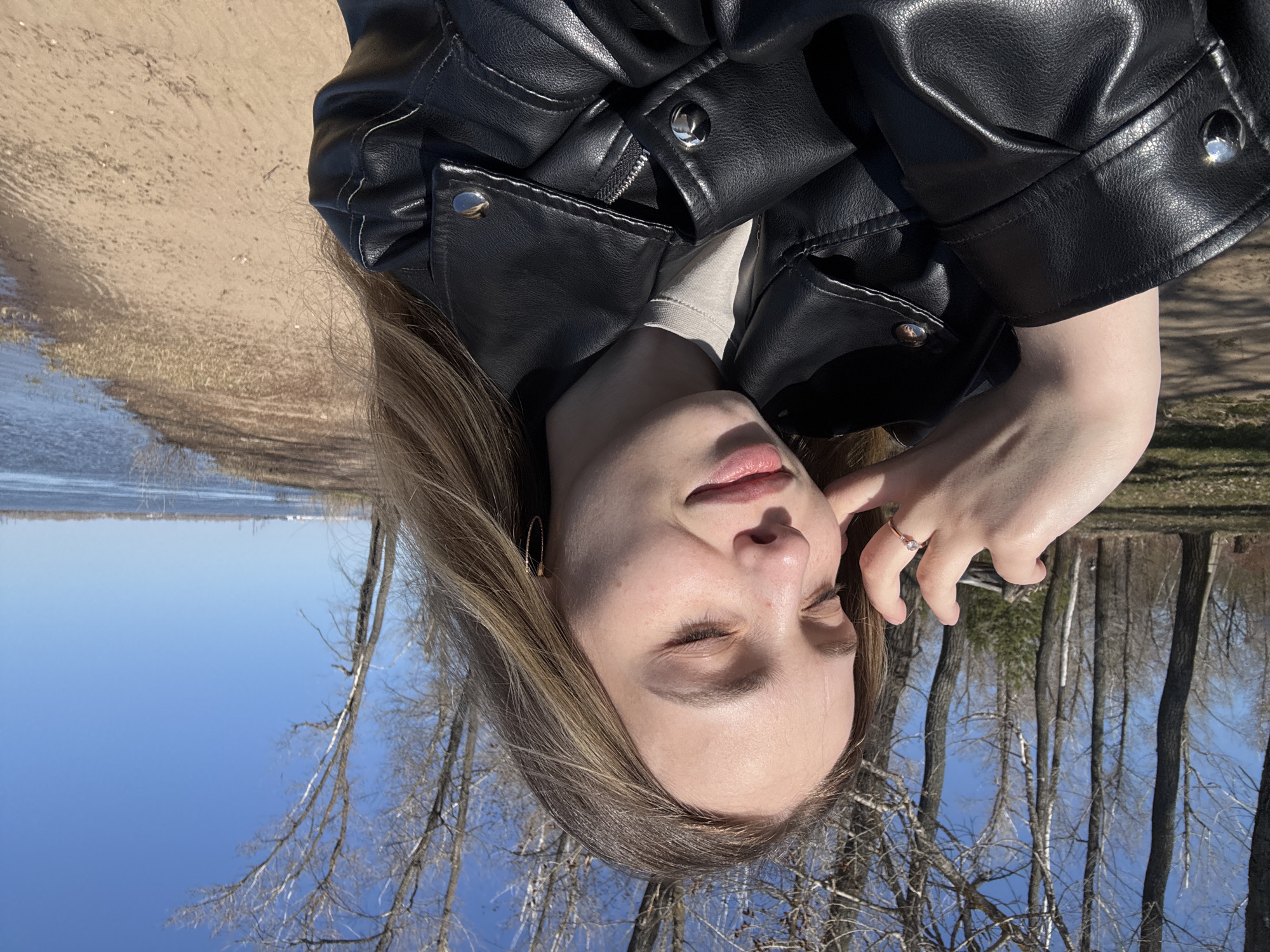 A woman wearing a black leather jacket stands outdoors. The background shows a body of water, sand, and trees without leaves. The image may have been taken near the city of Noviy, Russia.