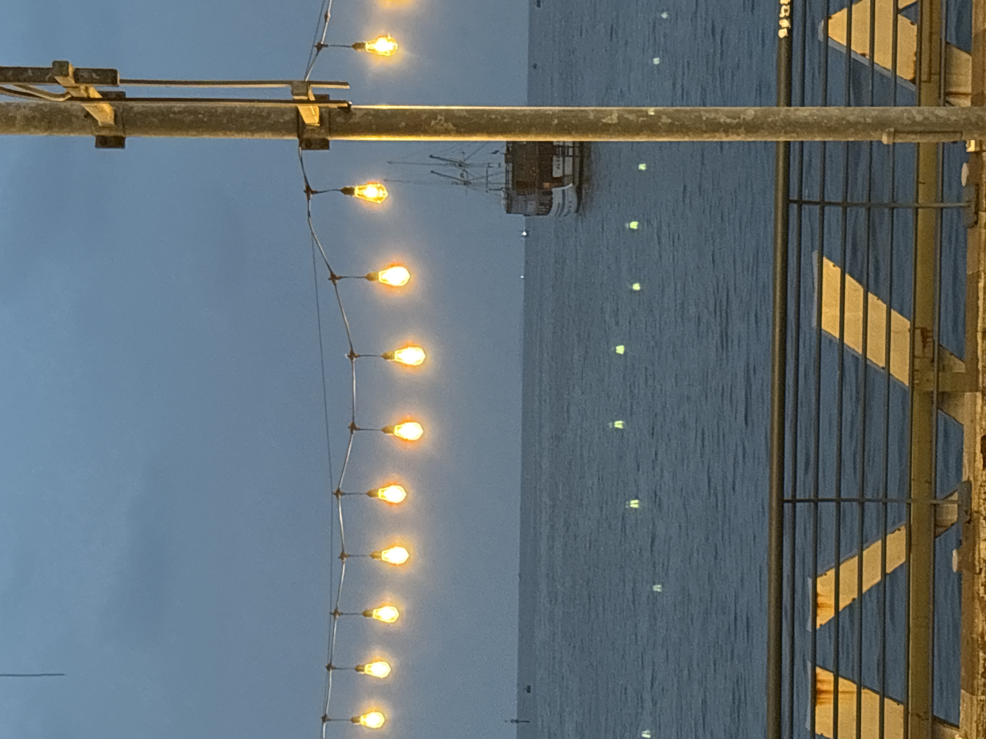 An eye-level outdoor shot shows the sea at dusk, viewed between string lights, a metal railing and metal support. The string lights are visible on the left side. The gray water is on the right and is partially reflecting lights. A fishing boat is in the distance. The scene appears to be located in North Jakarta, Indonesia.