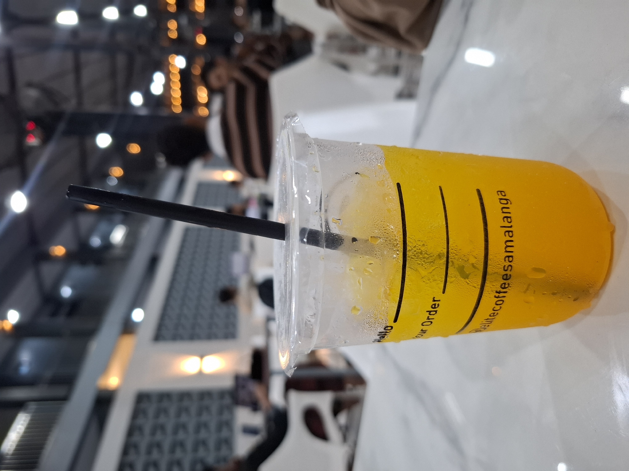 The image shows a close-up of a clear plastic cup filled with a bright orange beverage and ice, with a black straw. The cup has black text printed on it, with the words "Hello", "Your Order", and "delitecoffeesamalanga" visible. The cup is sweating with condensation. It is placed on a white marble-like surface. In the blurred background, several people are seated at tables in what appears to be a cafe or restaurant. The environment is dimly lit with warm, ambient lighting from overhead fixtures and wall sconces. The overall scene suggests a casual indoor setting, likely a cafe in Kampongbaro, Indonesia, given the text on the cup.