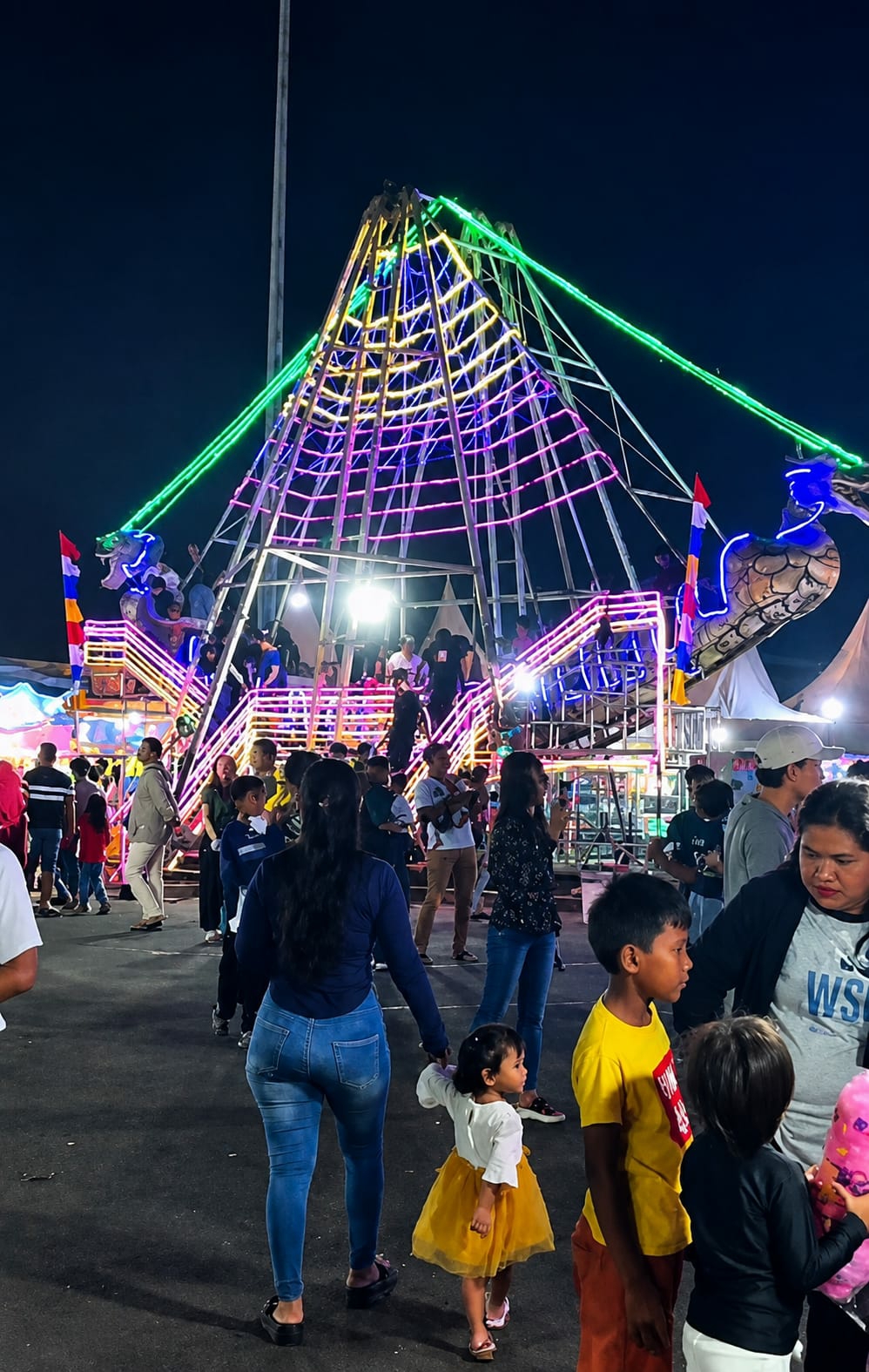 The image depicts a bustling nighttime scene at a fairground, possibly in the city of Medan, Indonesia, as indicated by the presence of flags and the general atmosphere. In the background, a large, pyramid-shaped amusement ride is illuminated by a vibrant display of green, purple, and yellow lights. The ride appears to be a pendulum or swing ride, with several gondolas visible. People are gathered around the ride, some waiting in line, others watching the ride in motion.
In the foreground and midground, a diverse crowd of people of various ages and ethnicities are milling about. Many are casually dressed, suggesting a relaxed, festive environment. A woman in blue jeans and a dark top is holding the hand of a young child wearing a yellow dress. Other individuals are seen walking, talking, or holding bags. The ground is paved, and the lighting from the rides and surrounding stalls creates a lively, somewhat chaotic ambiance. The overall impression is one of a popular public event, likely an attraction within the city of Medan.