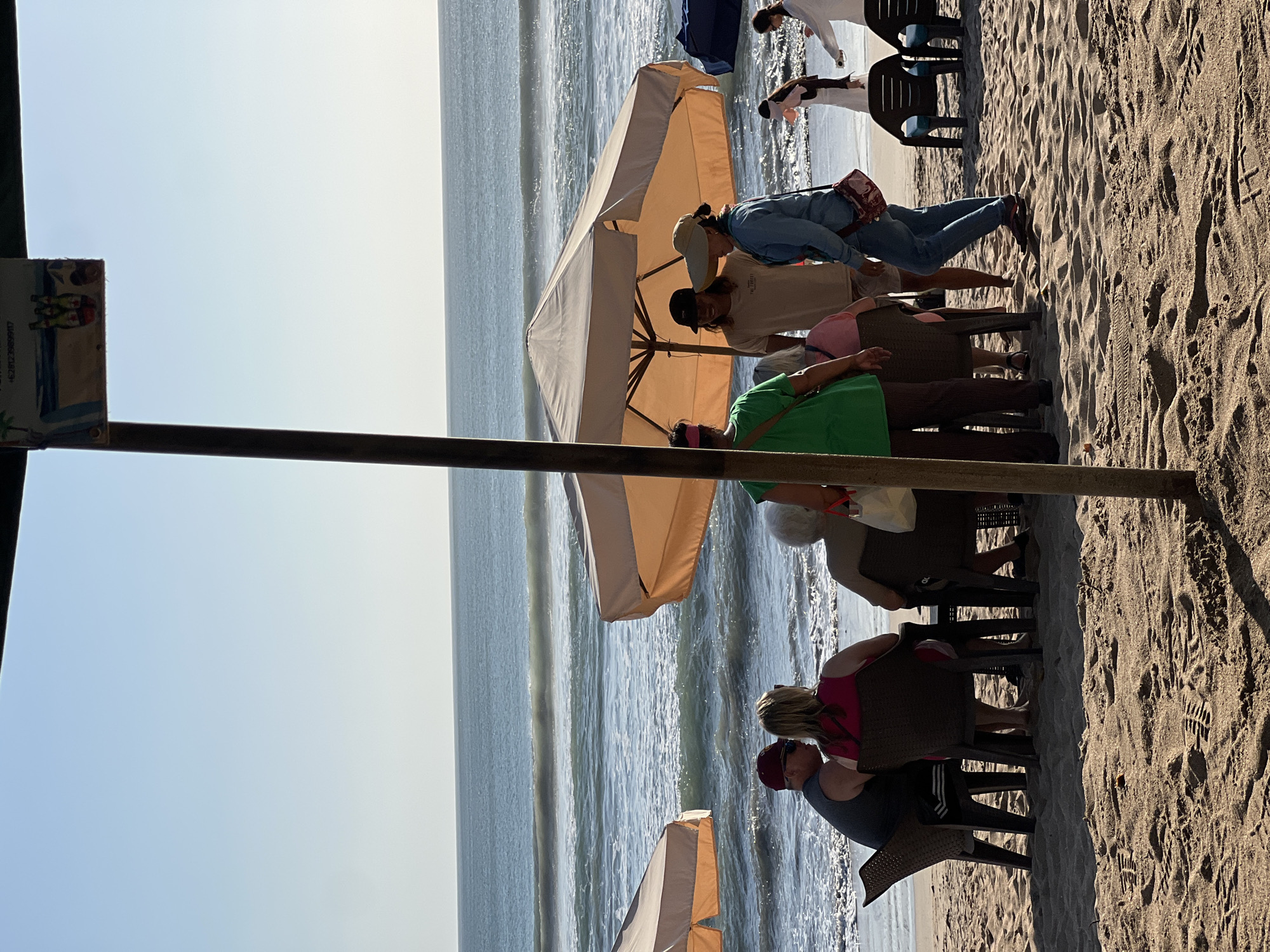 The image captures a beach scene in Kuta, Indonesia, with several people gathered under beach umbrellas. In the foreground, two people are seated at a table, facing away from the camera, with a third person seated beside them. To the right, a group of four people are standing, seemingly engaged in conversation, with one person holding a hat. Further in the background, two more individuals are walking along the beach. The environment is a sandy beach with the ocean and waves visible in the background under a clear blue sky. The lighting suggests it is daytime, likely late afternoon given the angle of the sun. Several beach chairs and umbrellas are scattered across the sand.