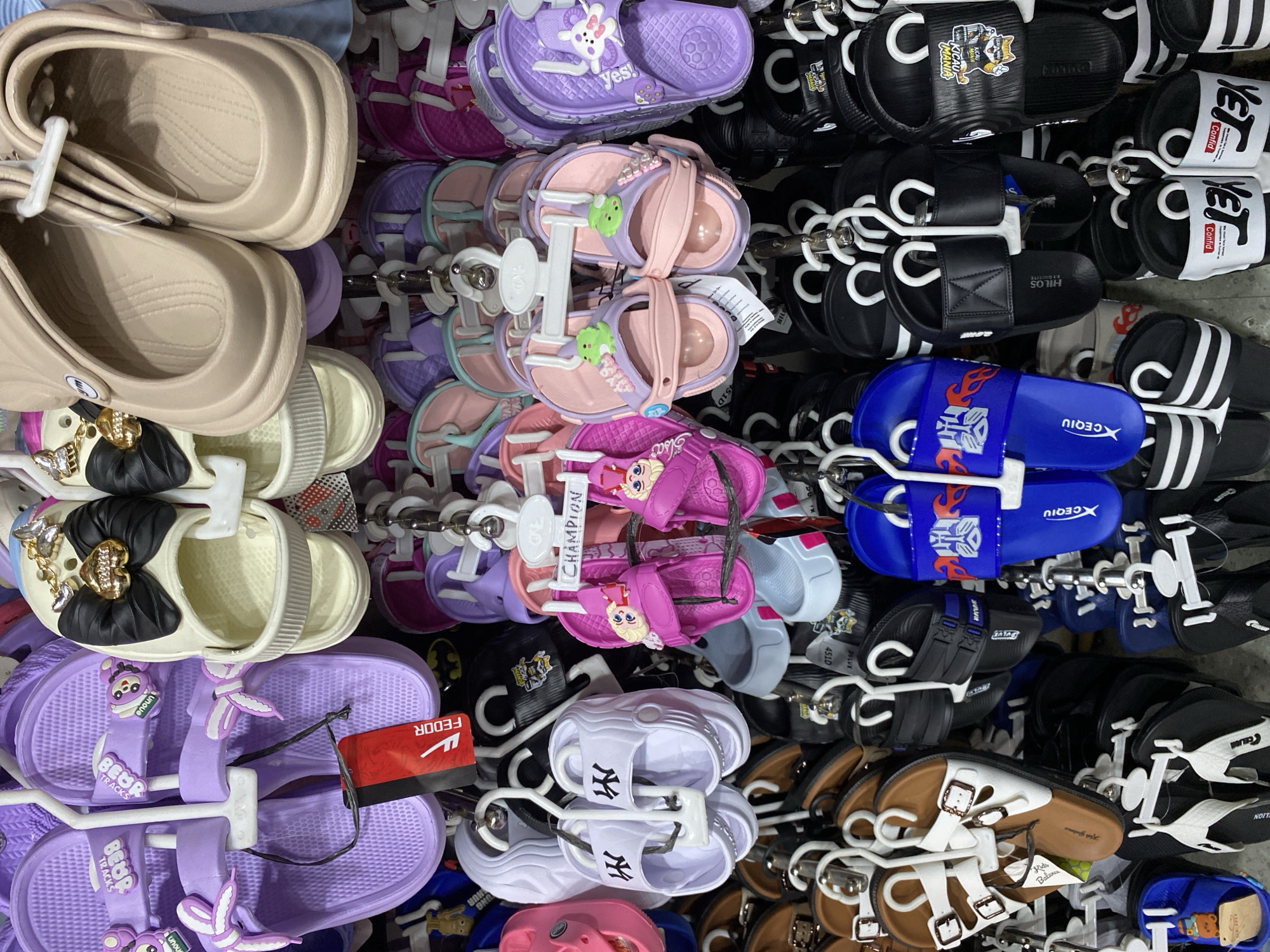 The image shows a retail display of various types of sandals and flip-flops for sale. The items are predominantly women's and children's footwear, featuring a range of colors and decorative elements such as bows, cartoon characters, and animal motifs. The footwear is hung from hooks and racks, suggesting an indoor market or store setting. The specific location mentioned, Garut, Indonesia, indicates this is a commercial establishment within that city. The image is a close-up view, focusing solely on the merchandise and the display fixtures. There are no people visible in the image.