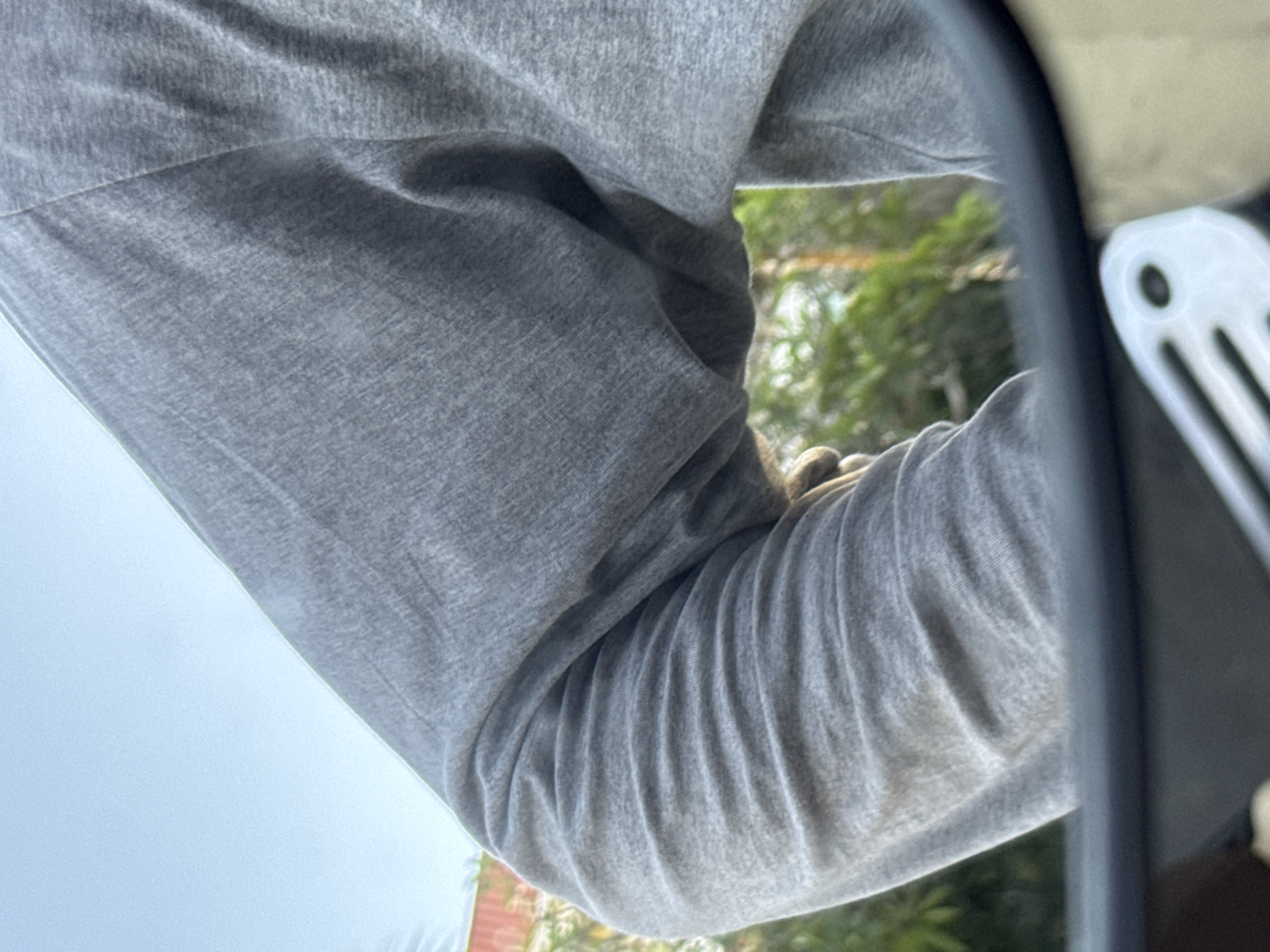 The image shows a close-up of a person's arm and shoulder, clad in a grey long-sleeved shirt. The fabric of the shirt is visible, with creases and folds suggesting movement. The arm is bent, with the forearm resting on something that is not fully visible but appears to be a light-colored surface. A portion of a mirror is reflected in the image, showing a blurred background of green foliage and some hints of what might be buildings or structures. On the right side of the frame, a white object with dark circular details is visible, possibly part of a vehicle or equipment. The overall lighting suggests it is daytime, with natural light illuminating the scene. There are no visible text elements in the image. The context provided is Kuta Selatan, Indonesia.