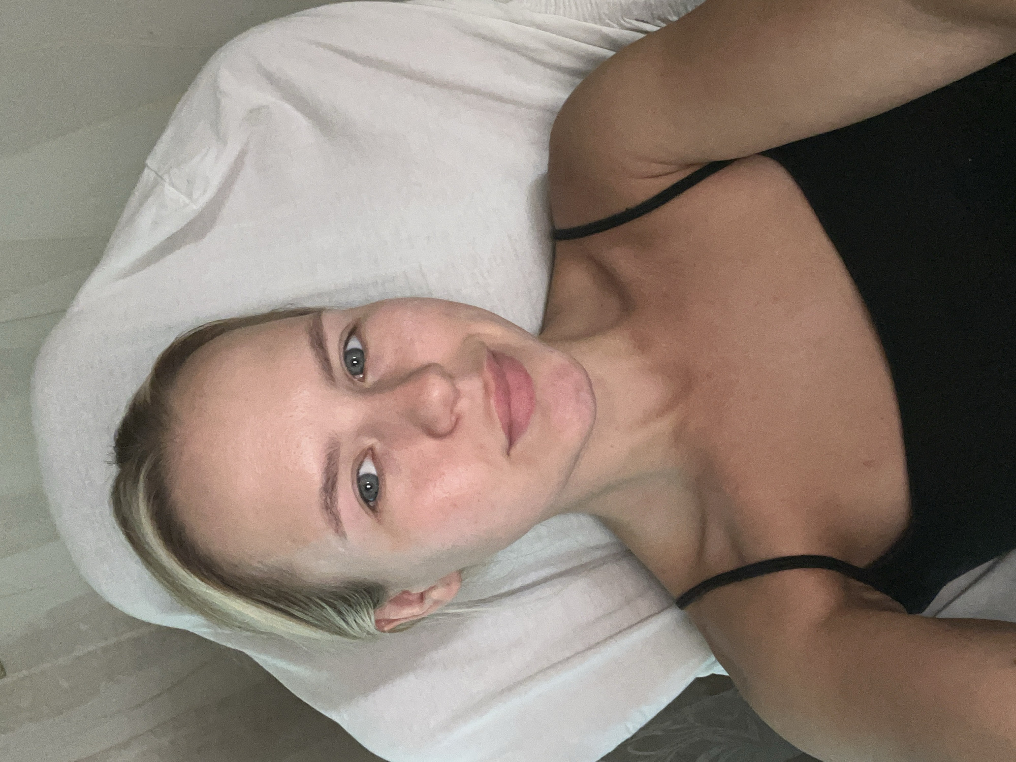 A young adult female with blonde hair is lying on a white surface, possibly a massage table or bed, in what appears to be an indoor setting. She is wearing a black spaghetti strap top. Her head is slightly tilted, and she is looking directly at the camera with a neutral expression. The background consists of a textured, light-colored wall or curtain and the white surface she is resting on. The lighting is soft and appears to be coming from above. The image is a selfie, taken from a high angle.