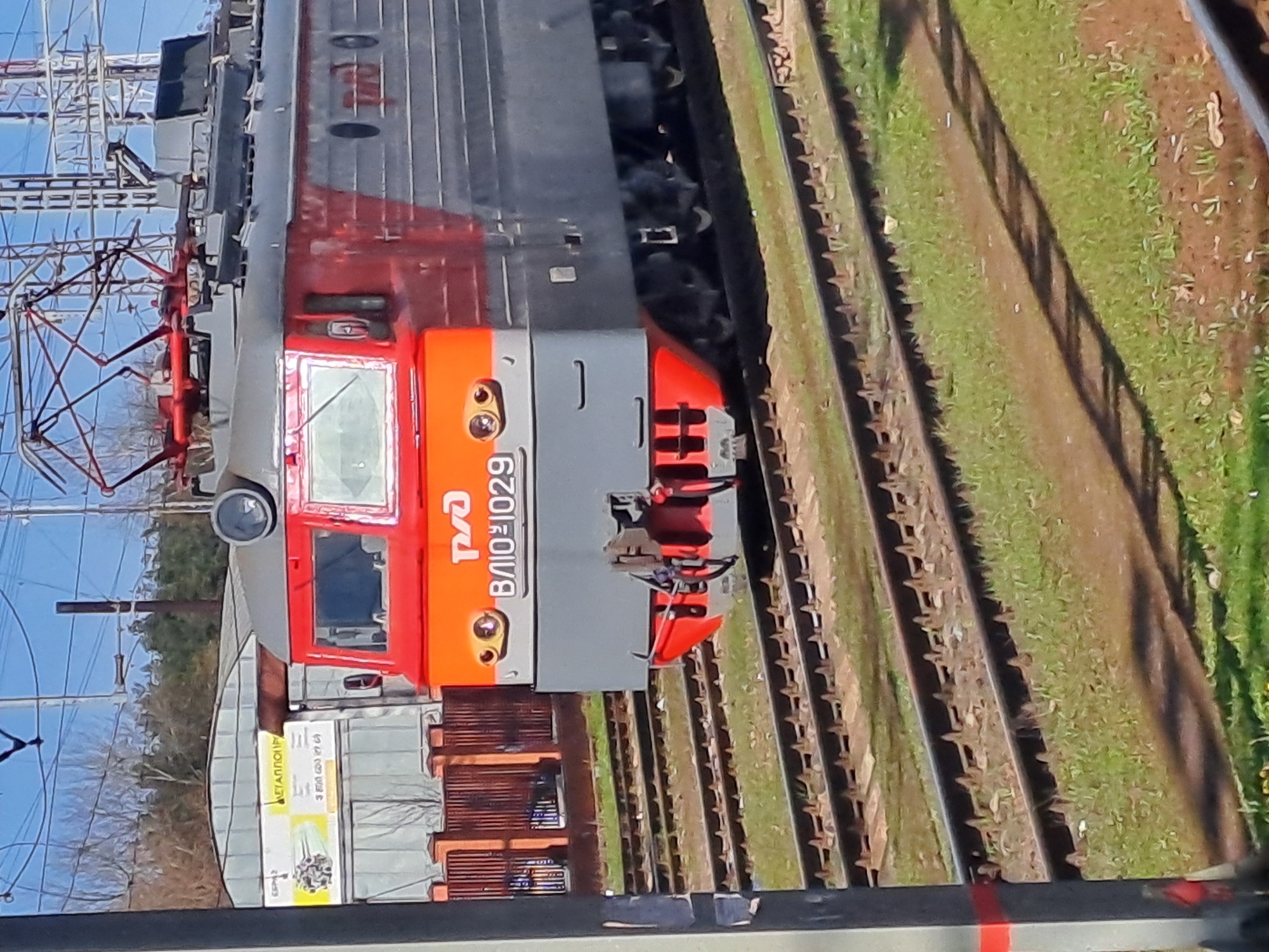 A close-up, angled shot depicts the front of a grey and red electric locomotive numbered VL10-1029. The locomotive is situated on a railway track, with greenery visible along the track's edge. Electrical power lines are overhead. A building with signs is partially visible in the background. The general context suggests the city of Fryazevo, Russia.