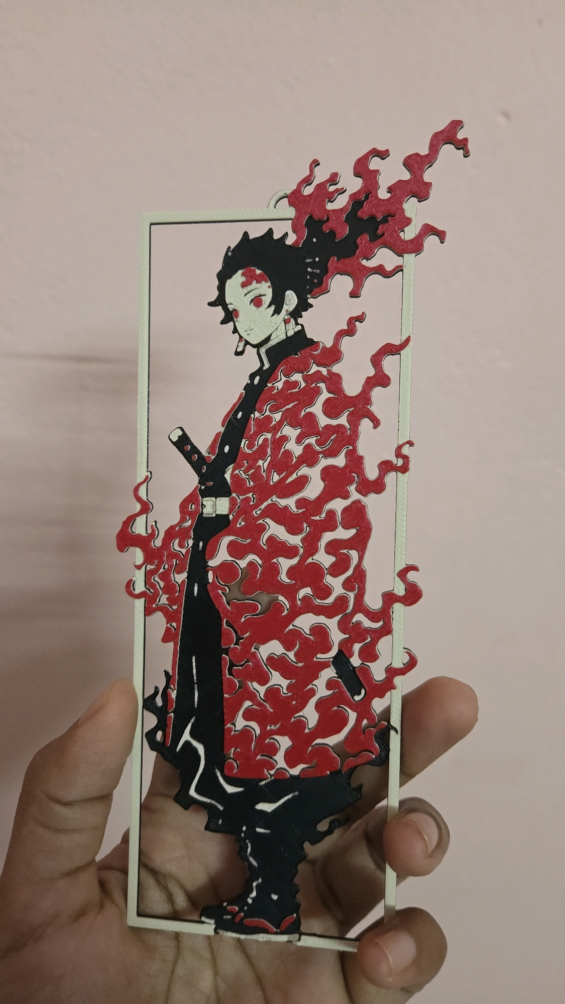 A vertical, full-body, cutout art piece of a Japanese anime character is held by a person's hand against a plain pink background. The character, depicted in a stylized, two-dimensional manner, has black hair with red flame-like accents, red eyes, and pale skin. They are wearing a black uniform with a white belt and a red, flame-patterned haori (a traditional Japanese jacket). A sword is sheathed at their hip. The cutout is framed by a thin, light-colored border. The background is a solid, pale pink color, typical of an indoor wall. The image is captured from a slightly downward angle, with the person's fingers visible at the bottom and right edges of the frame, suggesting they are holding the cutout at eye level. The lighting is soft and even, with no distinct shadows. There is no visible indication of the city of Chalakudy, India, in the image.