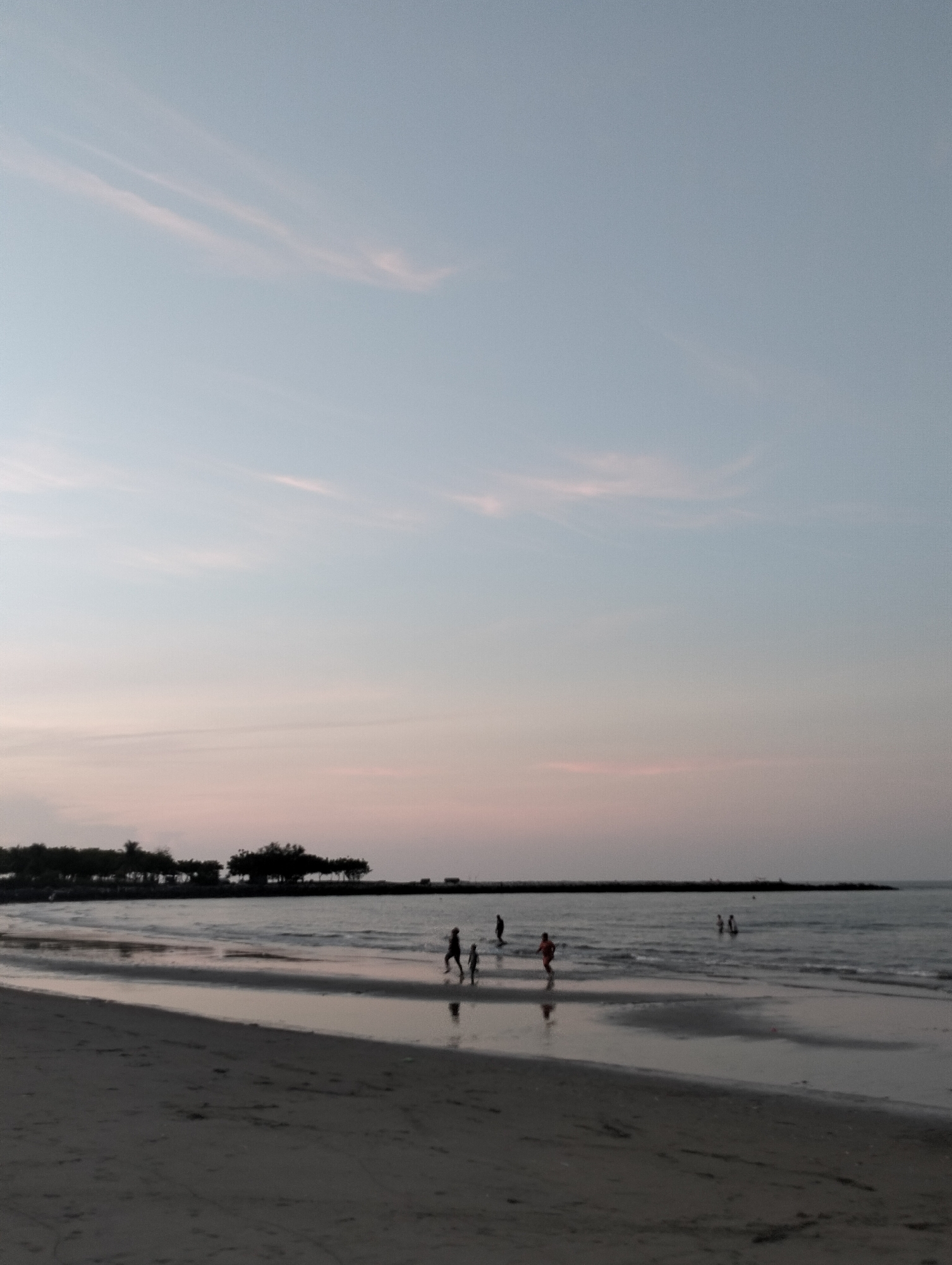 The image shows a beach scene in Lhokseumawe, Indonesia, at dusk. The sky is a pale blue with hints of pink. Several people are in the water, some standing and others appearing to be walking or running along the shoreline. The beach is a dark sand, reflecting the sky and the silhouettes of trees along the coastline. There is a dark structure or breakwater visible in the distance.