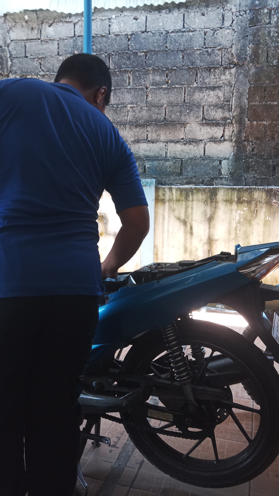 In an outdoor setting, likely a residential yard or alleyway in Jawa, Indonesia, a person is seen working on a light blue motorcycle. The individual, wearing a blue t-shirt and dark pants, is positioned with their back to the viewer and is leaning over the motorcycle, appearing to be engaged in some form of repair or maintenance. The motorcycle is supported by a kickstand and its rear wheel is visible. The background consists of a brick wall and a corrugated metal roof. The lighting suggests it is daytime.