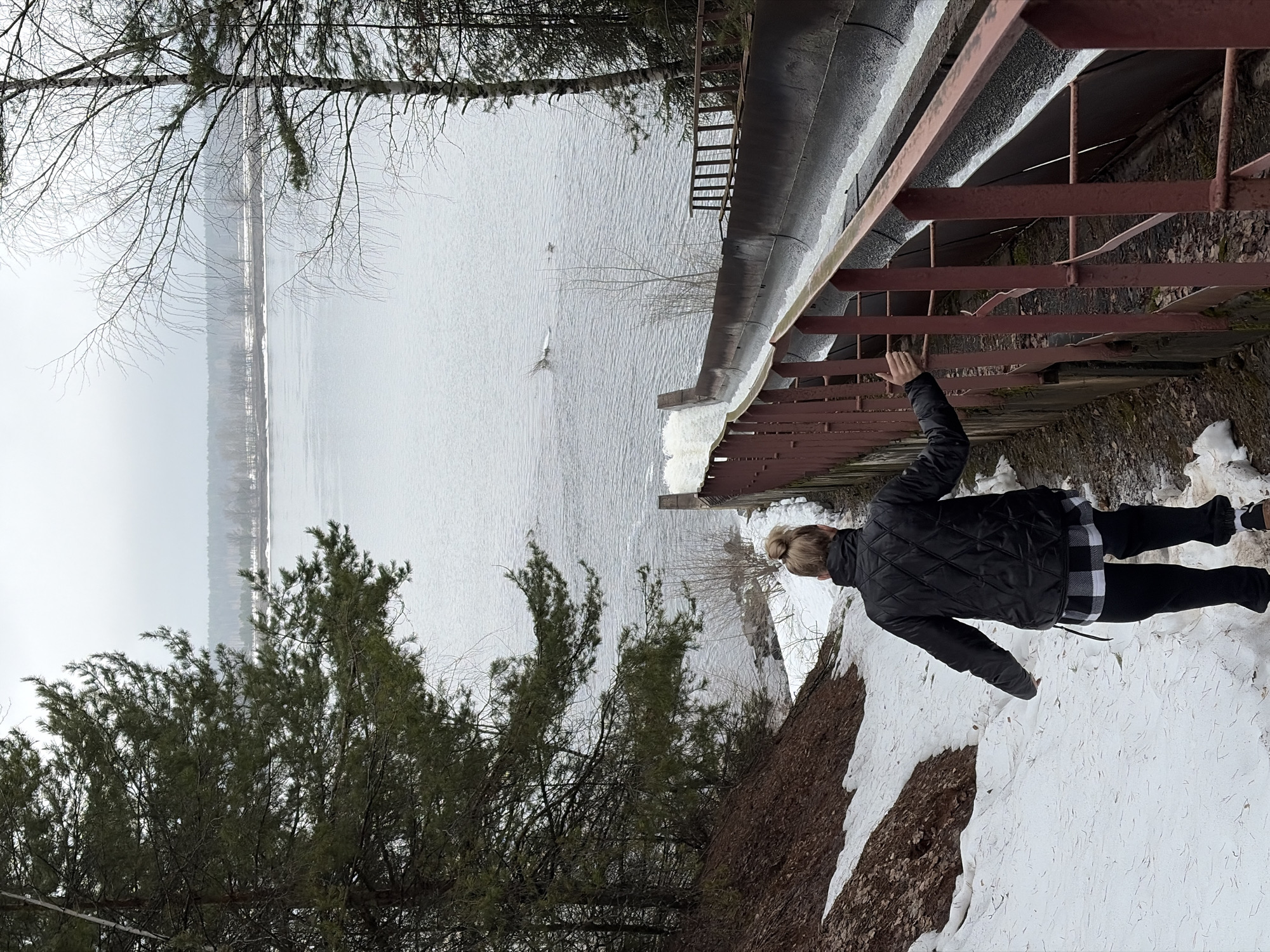 A woman stands on a snow-covered bank overlooking a body of water in Chaikovsky, Russia. The water is a greyish color, reflecting the overcast sky. Bare trees and evergreens line the far shore and the edge of the bank near the woman. A metal structure, possibly a large pipe with a ladder attached, runs along the edge of the bank. The woman is wearing a black quilted jacket, black pants, and brown shoes, with her hair in a bun. She extends one arm toward the metal structure. The scene appears to be outdoors during a cold season.