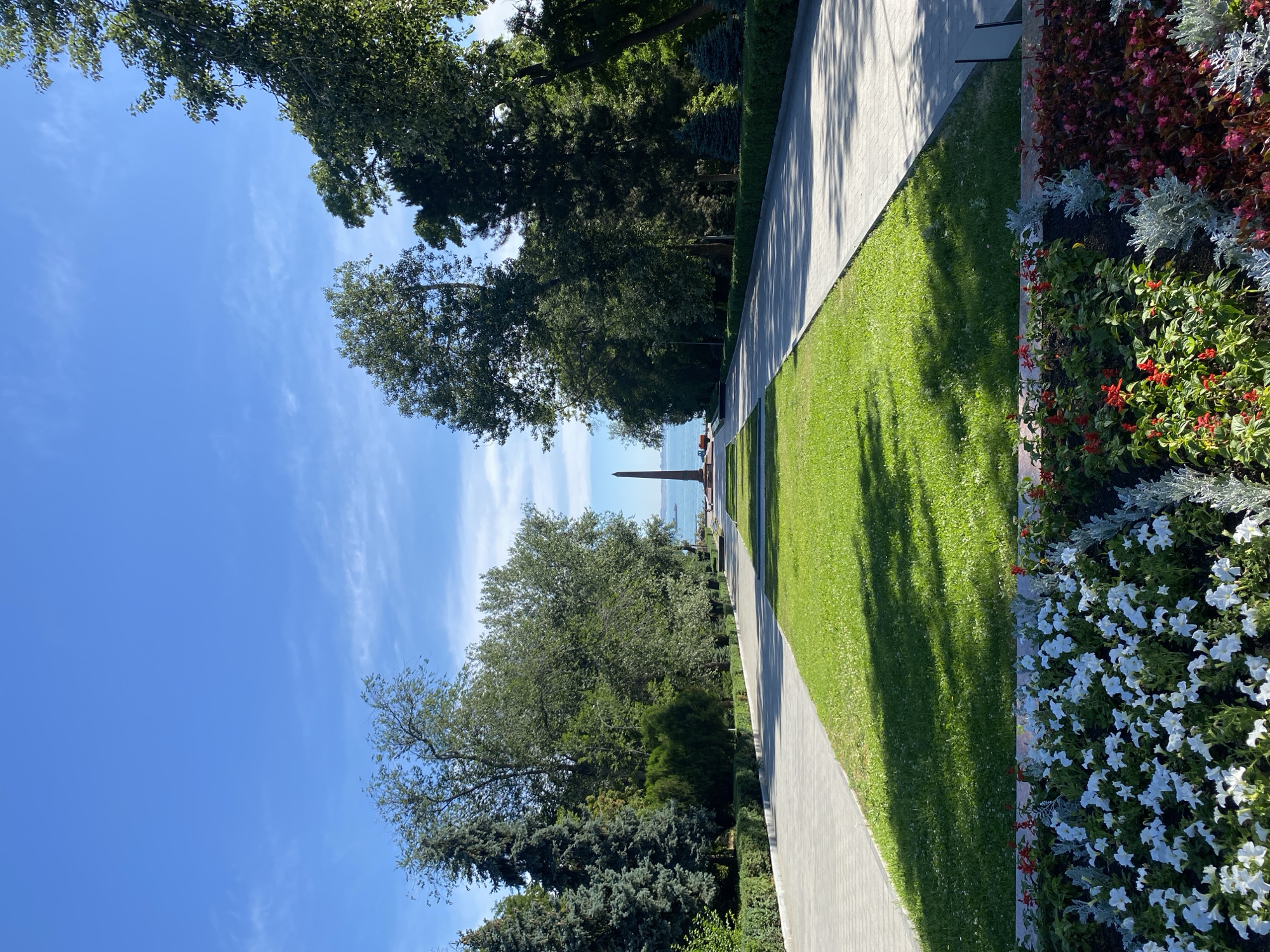An eye-level, vertical outdoor shot captures a symmetrical park pathway lined with lush green grass and vibrant flower beds in the foreground. Tall trees flank the path, leading the eye towards a distant obelisk monument standing tall against a clear blue sky with scattered wispy clouds. The obelisk appears to be situated near a body of water, likely a sea or large lake, indicated by a faint horizon line. The pathway is paved with light-colored interlocking stones and shows dappled sunlight and shadow from the overhead foliage. The flower beds at the bottom of the frame are a riot of color, featuring white, red, and pink blossoms, interspersed with silver-leafed plants. The scene evokes a sense of peace and grandeur within a well-maintained public space.