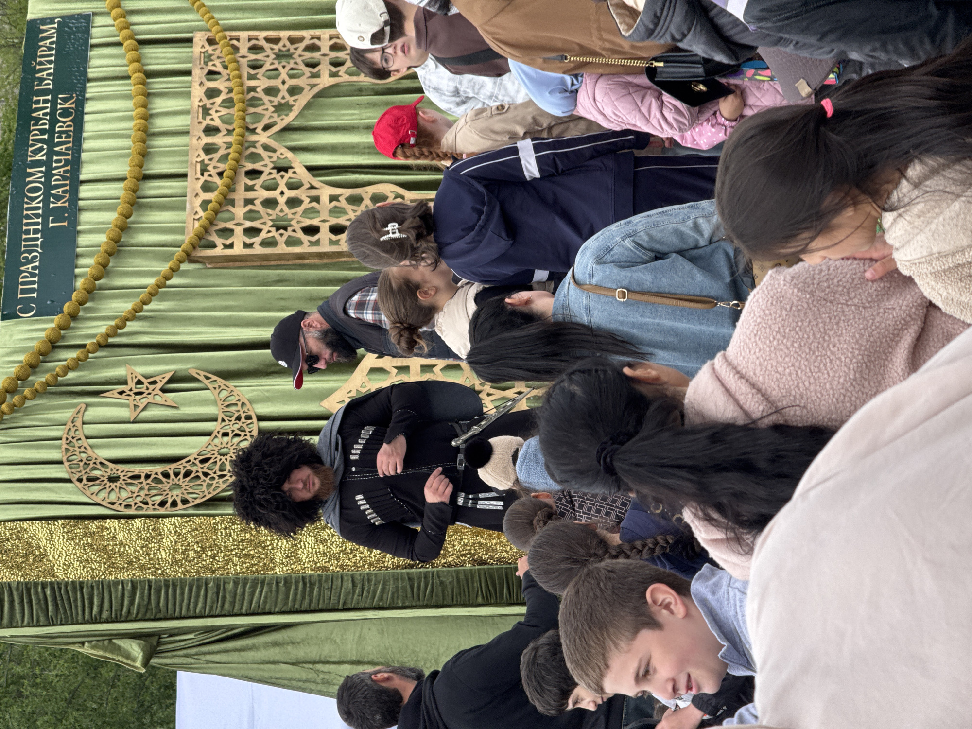 A group of people are gathered in front of a decorated display. One person in the display wears a black Cossack coat and a bushy black hat. Behind the person is a green backdrop decorated with a golden crescent moon and star, gold garlands, and a sign in Cyrillic text that mentions "Курбан Байрам, Г. КАРАЧАЕВСК". The sign indicates the location as Karachayevsk, Russia, and the religious holiday Kurban Bayram. People in the foreground are looking down, seemingly at something on a table or ground level.