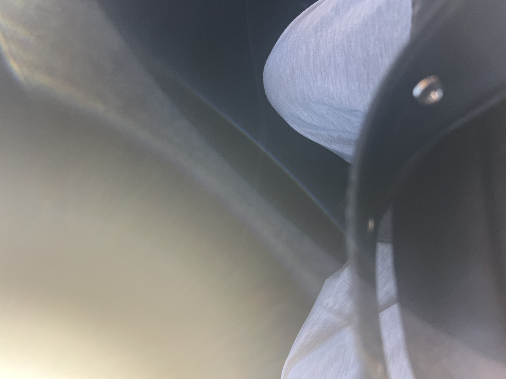 The media file is a close-up, abstract shot of what appears to be a person wearing white clothing, possibly a t-shirt or sweater, and a dark object, possibly a bag or part of clothing with a metal rivet. The lighting creates a lens flare effect, casting diagonal beams of light across the frame. A faint rainbow-like spectrum is visible within one of these light beams. The background is dark and indistinct. The focus is soft and the overall impression is impressionistic, with textures of fabric being the most discernible detail. The scene's setting or activity is unclear due to the abstract nature of the image. No text is visible.