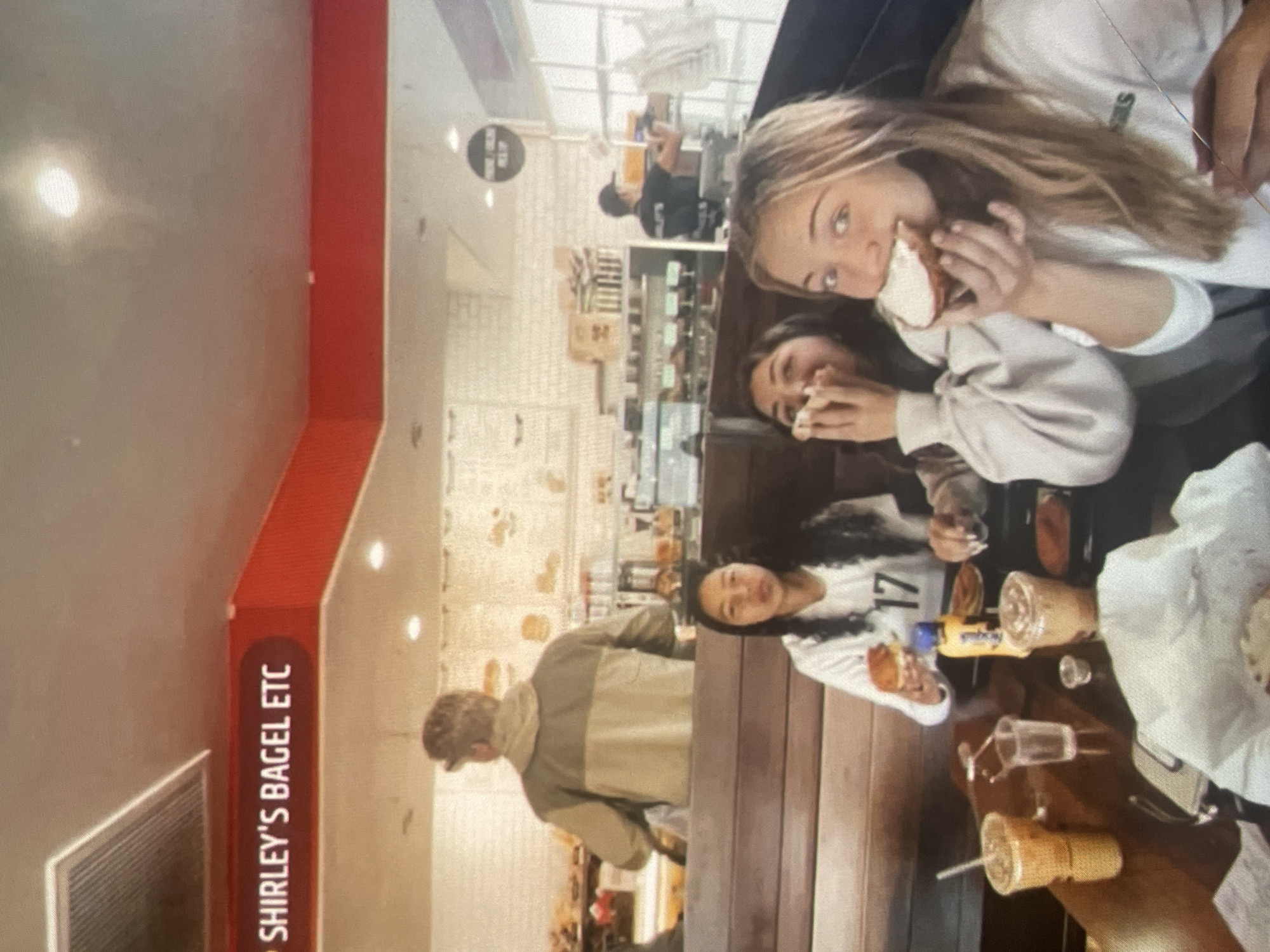 This media file captures an indoor scene at "SHIRLEY'S BAGEL ETC" in Harmanli, Bulgaria, likely a bagel shop or cafe. The image appears to be a photograph of a screen, resulting in some graininess.
The primary subjects are four individuals. In the foreground, two young women are seated at a wooden table, actively eating. One, with long blonde hair, is shown taking a bite out of a bagel or sandwich and looking towards the camera with a relaxed expression. The other, with darker hair, is also eating. Further back at the same table, another young woman with dark curly hair and a white top featuring the number "17" is looking towards the camera. A young man, wearing a two-tone jacket (khaki and light green/tan), stands in the middle ground with his back partially to the camera, appearing to wait or move within the establishment. Other blurry figures are visible in the background.
The activity taking place is people enjoying food and drinks in a casual setting. On the table, various objects are visible, including bagels, drinks in clear plastic cups with straws, a clear glass, and a small yellow container. The setting is bright, suggesting daytime, with light-colored walls, a red accent strip, and display counters in the background that hold various items. The overall atmosphere appears informal and social.
The most notable visible text is "SHIRLEY'S BAGEL ETC," displayed vertically on a red strip along the wall, serving as a clear identifier for the location. The number "17" is also visible on one woman's shirt.
