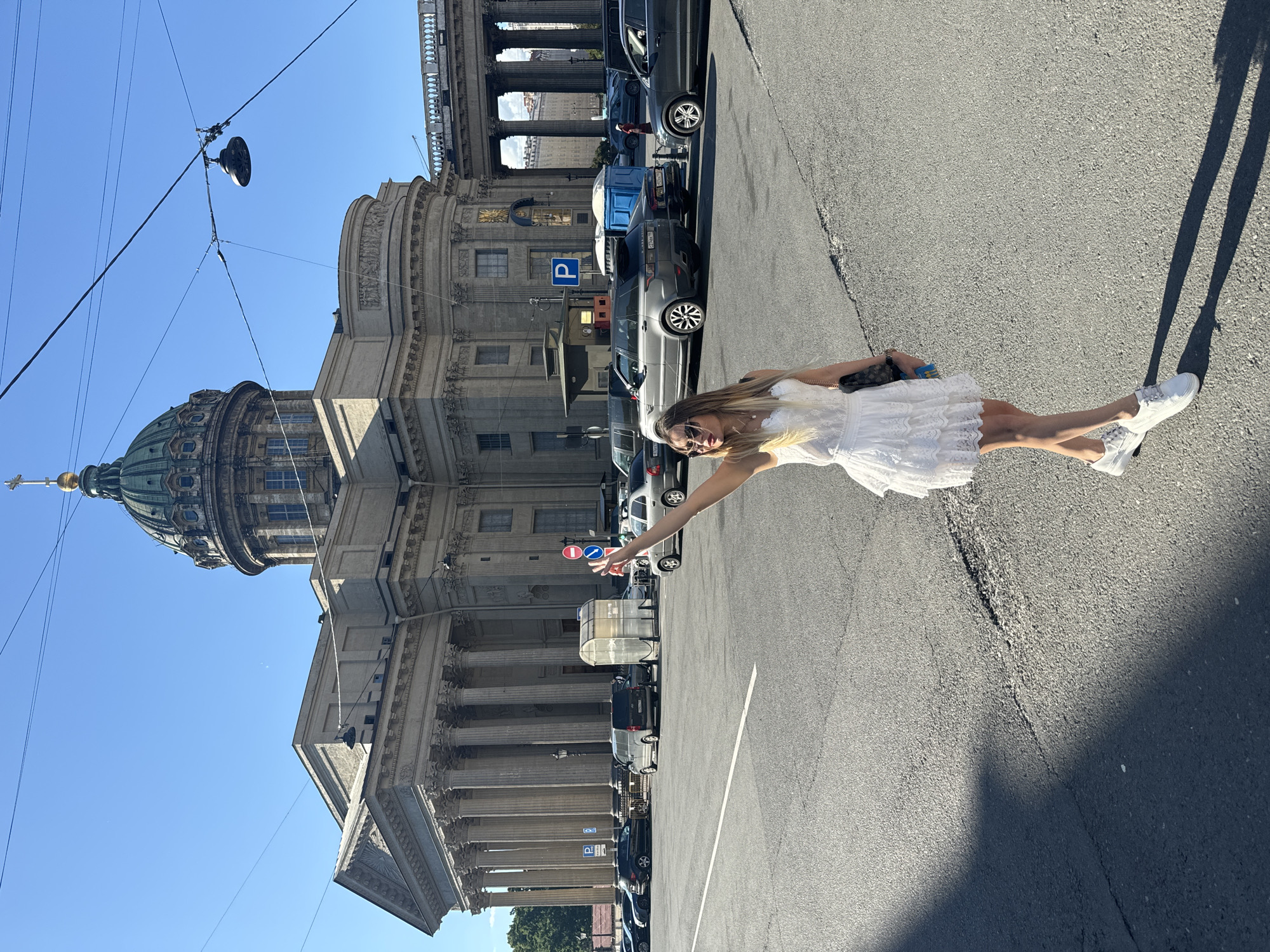 A young woman in a white lace dress and white sneakers is standing in a street in front of a grand building with a large dome. She is raising her right hand, palm facing forward, as if signaling or waving. She is wearing sunglasses and holding a dark bag in her left hand. The building behind her is the Kazan Cathedral, a prominent landmark in Saint Petersburg, Russia, recognizable by its semicircular colonnade and green dome. Several cars are parked on the street, and a portable toilet unit is visible to the right of the woman. The sky is clear and blue, suggesting a sunny day. The scene appears to be a street in the historical center of Saint Petersburg.