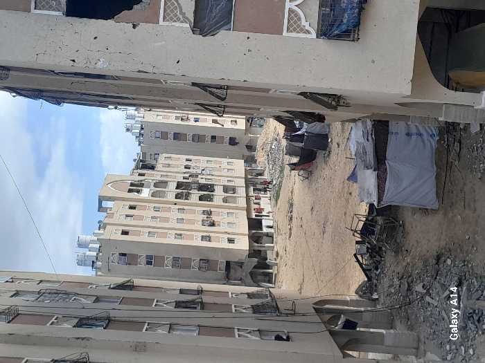 This image depicts a damaged residential complex in Khan Yunis, Palestinian Territory, under an overcast sky. The scene is dominated by multiple multi-story apartment buildings, constructed from light-colored concrete with brown trim and arched architectural details. Many windows on these buildings are broken, open, or covered with makeshift dark tarpaulins or fabric. The building on the immediate right exhibits extensive structural damage, including large missing sections of its facade and visible cracking. White cylindrical water tanks are visible on the rooftops of several buildings.
The ground between the buildings is an unpaved, sandy, and dusty area filled with debris, rubble, and discarded materials. In the midground, various makeshift shelters are present. One prominent shelter on the left is constructed from a metal frame and covered with a white tarp, partially displaying "UNICEF" branding. Other, smaller shelters made of dark fabric or tarpaulins are situated further back. Scattered across the ground are remnants of destroyed structures, including twisted metal frames and concrete fragments. In the far distance of the central courtyard, a small group of approximately four individuals, indistinct but appearing to be adults and possibly children, is visible walking or standing near the base of additional damaged buildings. A thin wire crosses the upper portion of the frame.