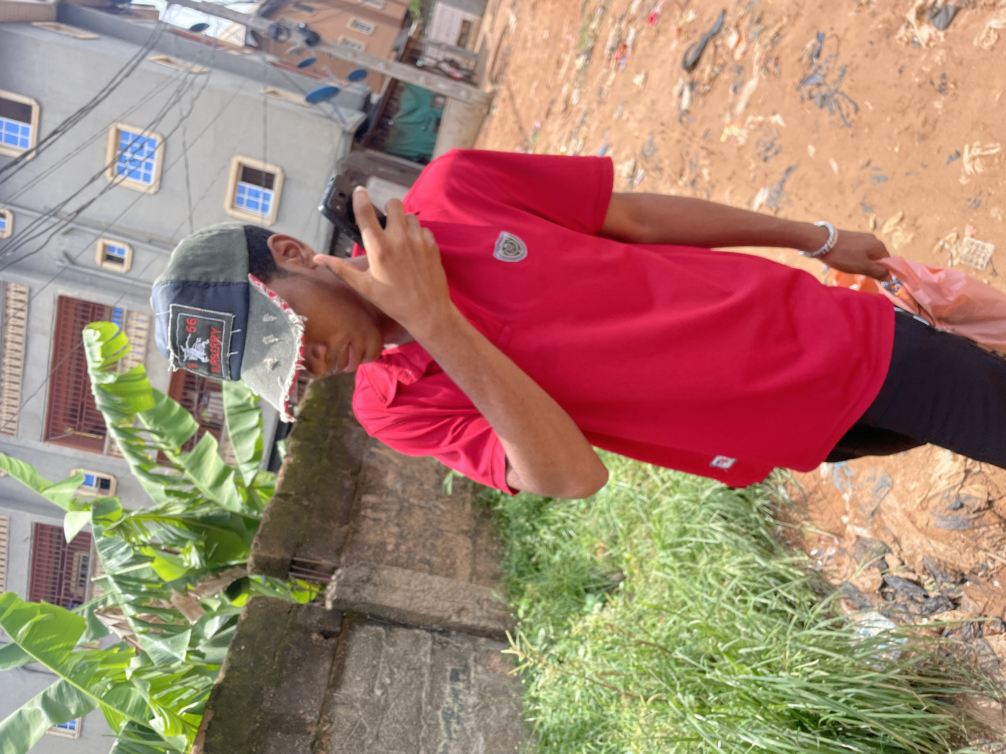 The image depicts a person in Nkpor, Nigeria, holding a phone to their ear. The individual is wearing a red t-shirt, black pants, and a baseball cap. They are standing on an uneven ground surface that transitions from overgrown green vegetation to bare ground littered with debris. In the background, there is a building with multiple windows and a banana tree next to a concrete fence. Power lines are visible above the building.