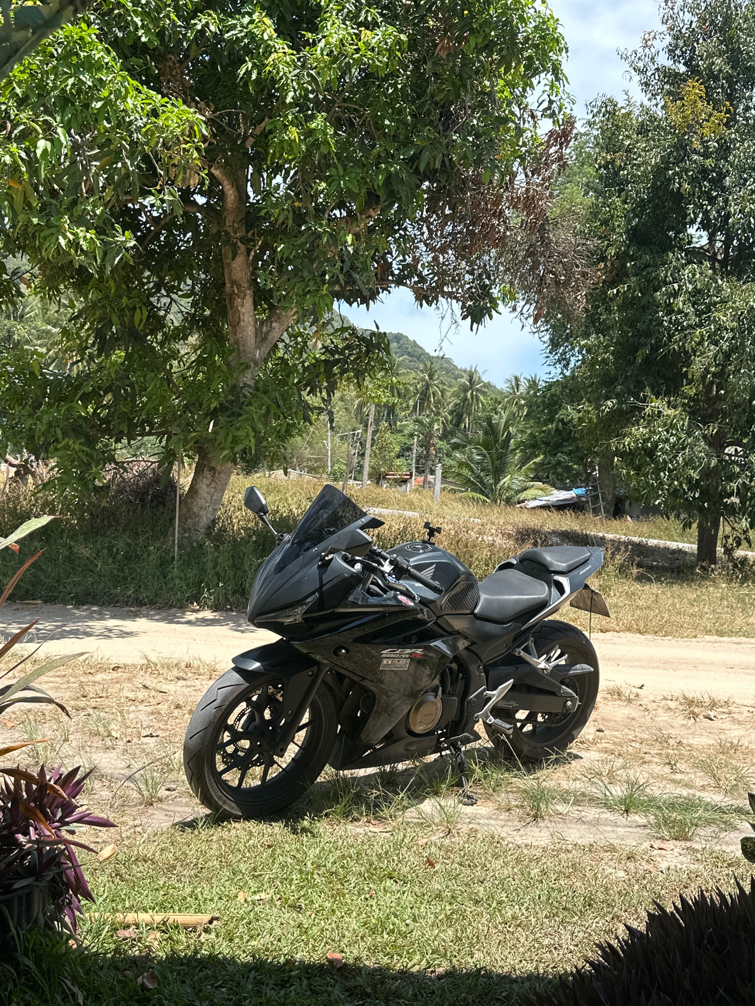 A black motorcycle is parked on a dirt path in front of a tropical landscape. The motorcycle is facing left and is angled slightly towards the viewer. It has a sleek, sporty design with a black windscreen and black seats. The background features lush green trees, palm trees, and a hazy sky. The ground is a mix of dirt and dry grass, with some green grass in the foreground. The scene appears to be a sunny day in a rural or tropical setting, possibly in Ko Pha-ngan District, Thailand, given the vegetation. There are no people visible in the image.