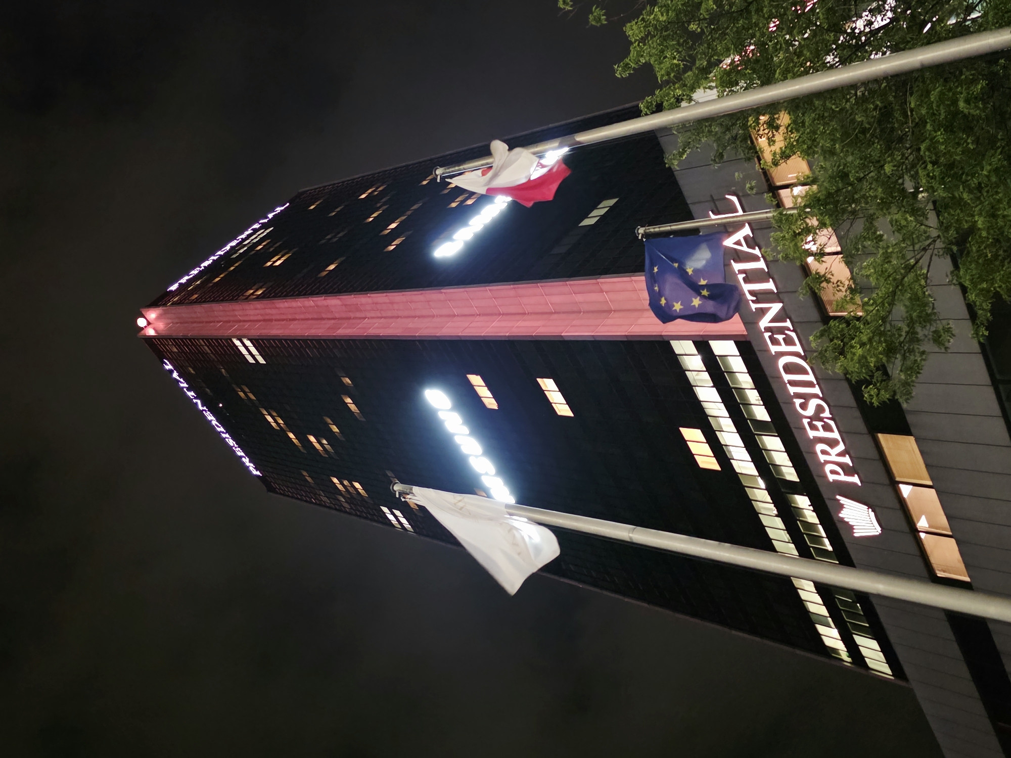 The image shows a low-angle, night-time view of the Presidential Building in Warsaw, Poland. The skyscraper is illuminated by its own lights and has "PRESIDENTIAL" written in large, glowing letters across its facade. A red vertical strip runs up the center of the building, and windows are lit from within. Two flags are visible on poles in front of the building: one is a white flag, and the other is a red and white flag, possibly the flag of Poland. A third flag, the flag of the European Union, is also visible. Green trees with lush foliage are in the foreground, partially obscuring the lower levels of the building. The sky is dark and overcast, with some clouds visible.
