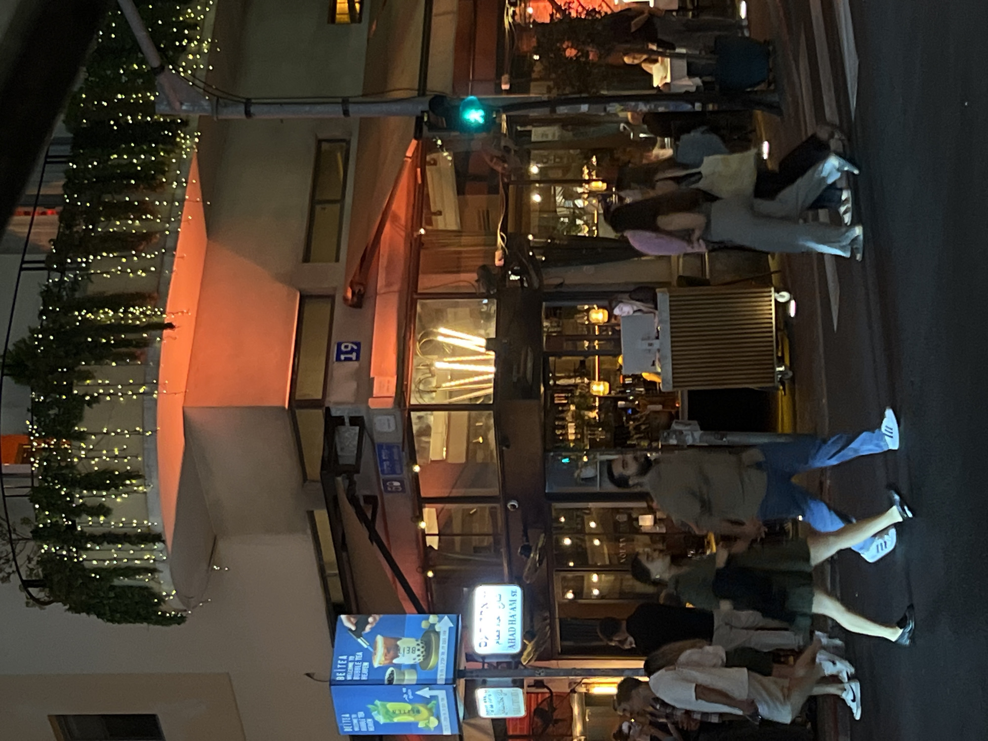 Here's a detailed description of the image:
**General Description:** The image appears to be taken at night in an urban setting. It is dominated by a multi-story building on the left side, and there are pedestrians on what looks like a crosswalk. The lighting is a mix of streetlights, lights from buildings, and festive lights on the building's facade.
**Location:** Based on the signage visible ("AHAD HA'AM ST." and some Hebrew script), this is likely Ahad Ha'Am Street in Tel Aviv, Israel.
**Building Details:**
* The building on the left is adorned with strings of small lights, possibly for decoration.
* There are windows and signs on the building. The number "19" is visible.
**People:**
* Several pedestrians are present, mostly walking on the crosswalk.
* The focus seems to be on a group of people crossing the street.
* The people are dressed in casual clothing.
**Objects:**
* There's a traffic light visible at the top of the image, with a green light showing.
* Signs for "BEITEA" (a bubble tea place), and Ahad Ha'Am st. sign are visible.
* There seem to be chairs and tables outside some establishments, indicating restaurants or cafes.
**Actions and Interactions:**
* People are walking, suggesting normal pedestrian activity.
* The green light indicates it is safe for pedestrians to cross.
**Environment:**
* It's night time.
* The lighting is relatively bright due to streetlights and building illumination.
* The scene is lively and bustling, typical of a commercial area.