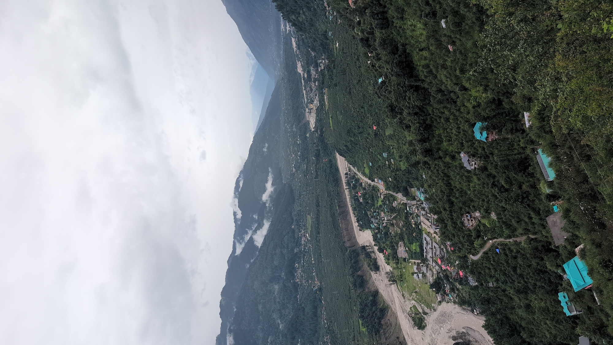 An aerial shot captures a verdant valley nestled between imposing mountains, under a blanket of overcast, grey skies. The valley floor is bisected by a wide, muddy river, its banks lined with lush vegetation and scattered human settlements. Buildings, some with vibrant turquoise roofs, are interspersed with the dense, dark green foliage of trees that cloak the lower slopes of the mountains. A winding road snakes through the valley, connecting the clusters of houses and a more densely populated area on the river's edge. The upper reaches of the mountains are shrouded in mist and clouds, creating a sense of depth and mystery. The scene suggests a remote, possibly mountainous region, characterized by its natural beauty and the harmonious coexistence of human habitation with the surrounding wilderness.