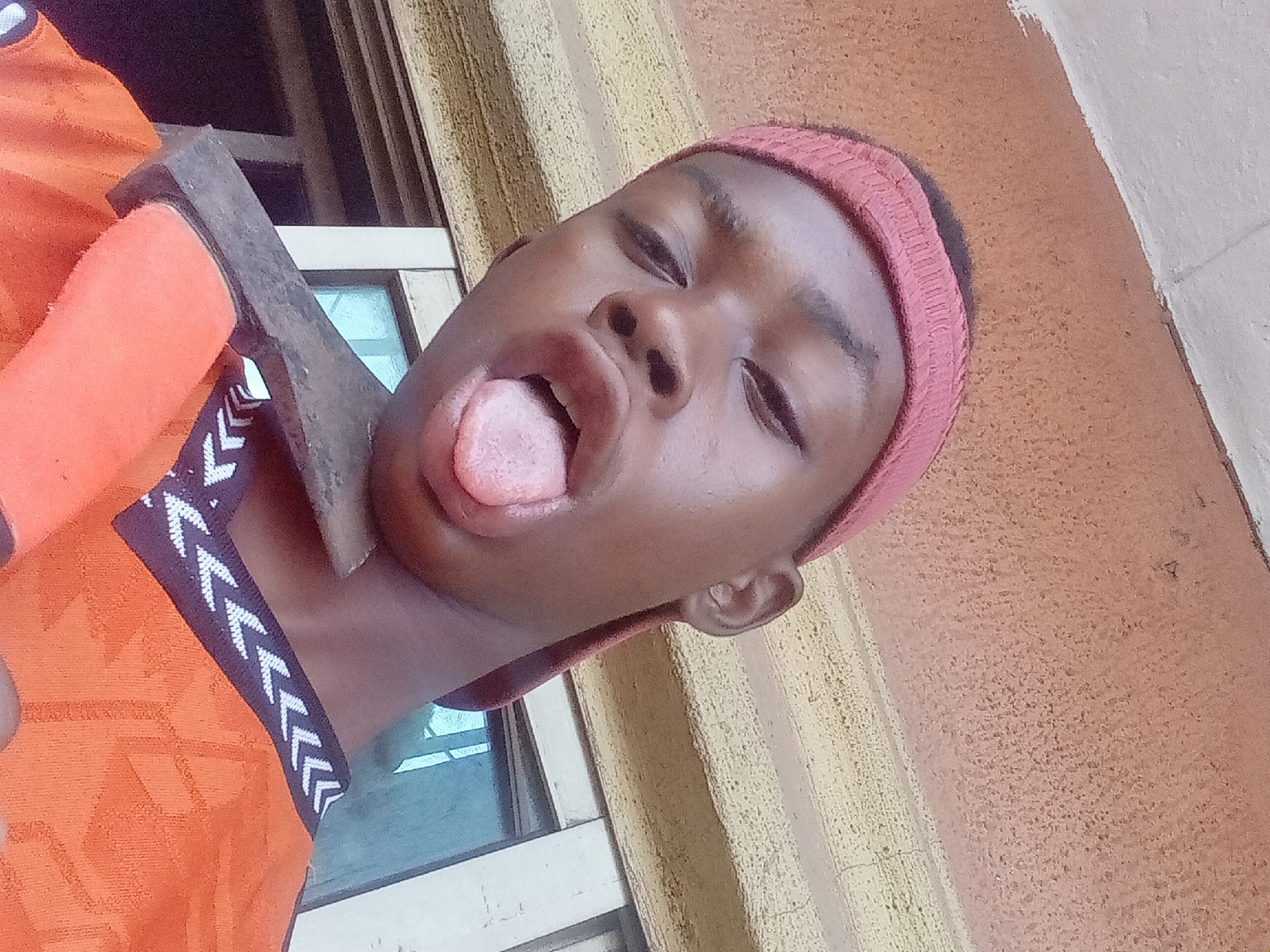 A young person is posing with an axe near a window. The young person, possibly male, has dark skin, a pink headband, and is wearing an orange shirt with white and dark trim. They are sticking out their tongue. An axe with an orange handle and a gray head is positioned near their neck. The background includes a window and a wall with textured paint in beige and orange tones. The location is presumably Nkpor, Nigeria.