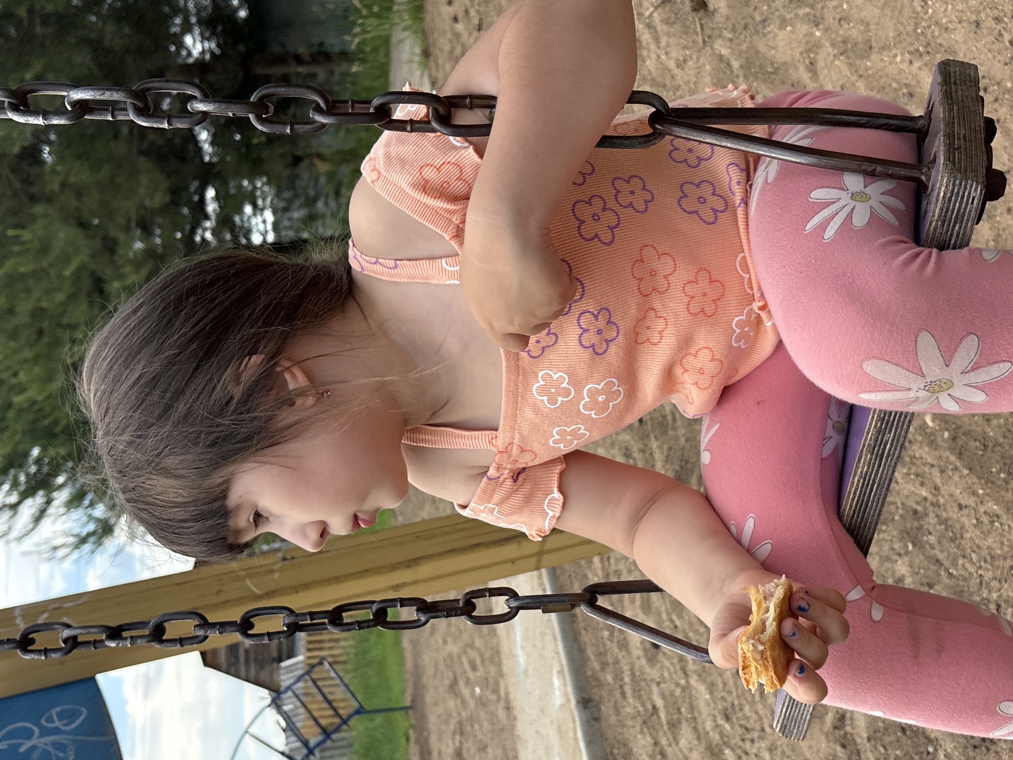 A young girl sits on a swing with a small piece of food in her hand. She is wearing a peach-colored, off-the-shoulder shirt with a floral pattern and pink leggings with a white floral design. The swing set has thick metal chains and a wooden seat. The background shows a playground area with sand, a wooden structure, and trees. The scene appears to be outdoors during the daytime. The location is the city of Chaikovsky, Russia.