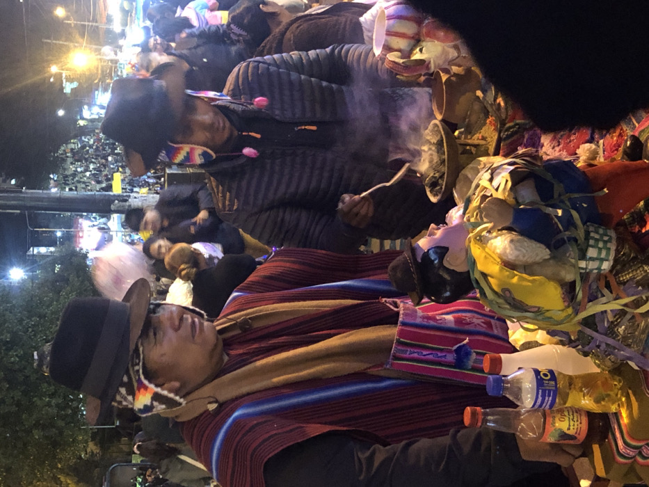 A person wearing a hat and colorful shawl is lying down, possibly deceased, surrounded by bottles and other objects. Beside them, a puppet or doll with a hat lies in a similar position. Smoke rises from a bowl held by another individual, who is bundled in a thick jacket. In the background, a crowd is visible, suggesting a public event or procession, possibly with religious or cultural significance. The scene is dimly lit, casting shadows and creating a somber atmosphere.