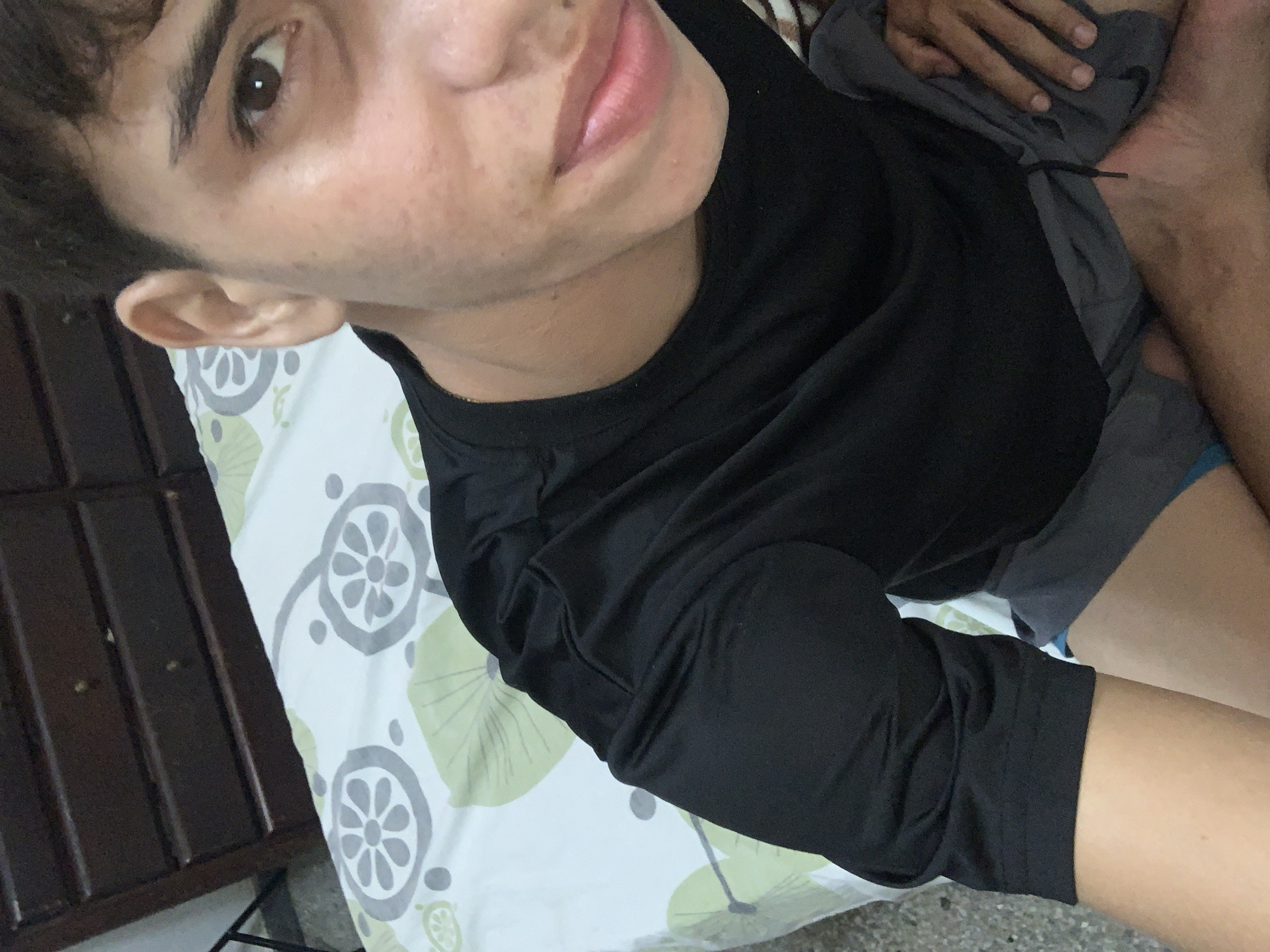 A person wearing a black t-shirt and gray shorts is lying on a bed with a white patterned bedspread. The person has short dark hair and light skin. The bed frame is visible on the left side of the frame. The environment appears to be a bedroom. There is no information to indicate the location is in El Valle del Espíritu Santo, Venezuela.