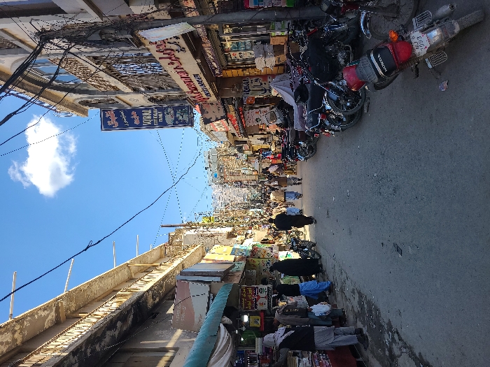 The image portrays a bustling commercial street in Quetta City Tehsil, Pakistan, under a clear blue sky featuring a single cumulus cloud. The narrow, paved thoroughfare is bordered on both sides by multi-story buildings housing shops and market stalls. A dense network of black electrical wires is suspended overhead, crisscrossing between utility poles.
On the left, a market stall operates under a green awning, displaying various goods. Several pedestrians are present on this side, including two individuals attired in black abayas. On the right, a line of motorcycles is parked along the sidewalk, with a red Honda motorcycle prominently positioned in the foreground, displaying a partial license plate (MJ 7003). Shopfronts on the right feature displayed merchandise and vertical signage, including text in Urdu script and "NATIONAL SOBHAN HOTEL FAMILY."
Approximately twenty individuals are visible walking along the street, primarily moving away from the camera's perspective. Most are dressed in traditional attire. The street surface is asphalt, exhibiting wear and scattered minor debris.