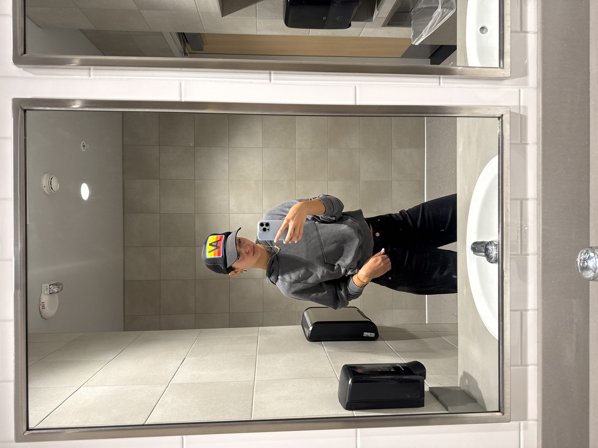 The image shows a mirror selfie taken in a bathroom in Los Angeles, United States of America. The person in the reflection is wearing a gray hoodie, black pants, and a baseball cap with a colorful logo. They are holding a smartphone. The bathroom has tiled walls and floors, with a sink, paper towel dispensers, and other bathroom fixtures visible in the reflection. The mirror has a metallic frame.