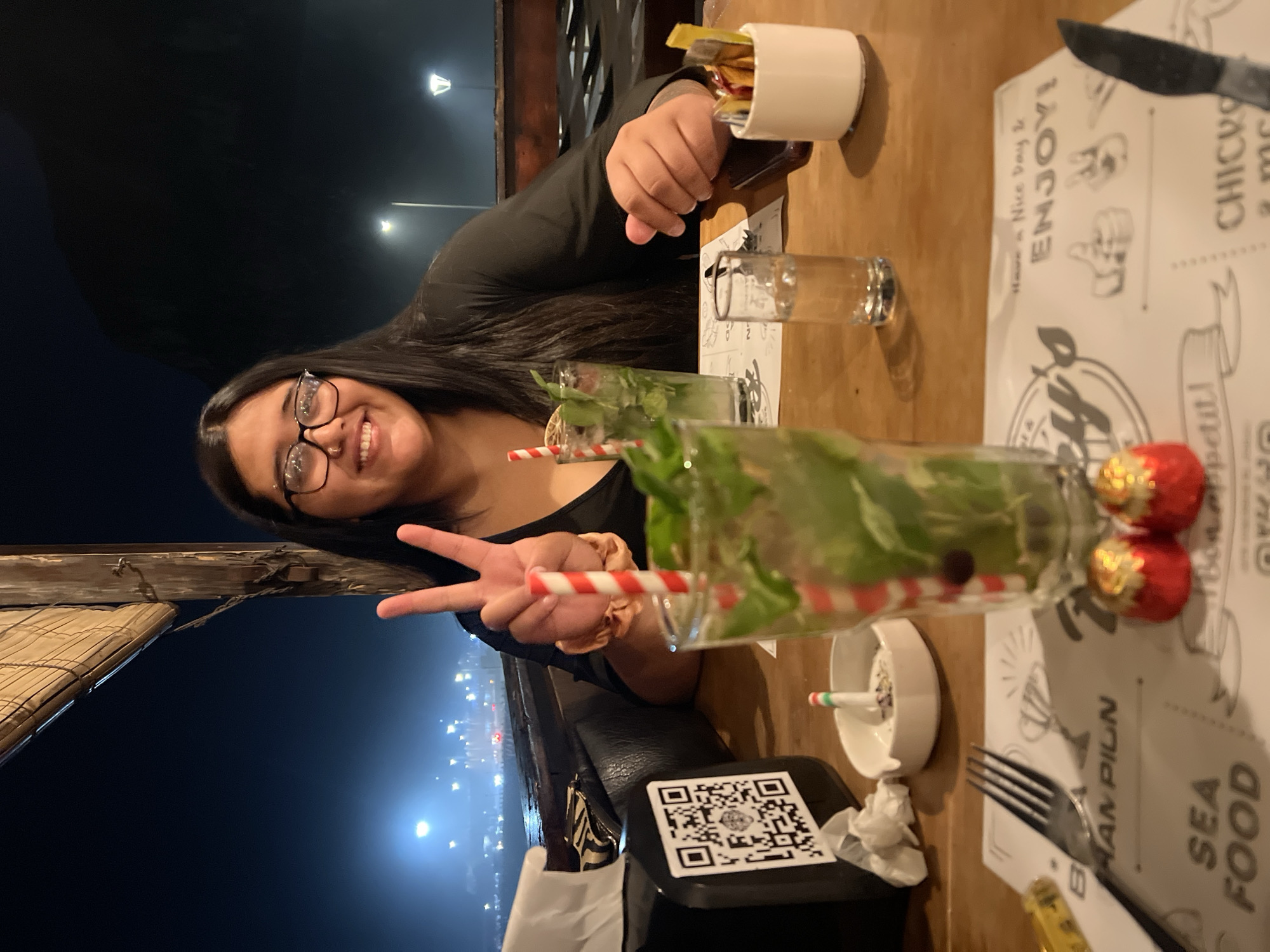 This nighttime image captures a young woman seated at a wooden table in a semi-outdoor restaurant setting, offering a view of a coastal city. The woman, with long dark hair and eyeglasses, smiles directly at the camera. Her right hand is raised, forming a peace sign, with a light-colored scrunchie visible on her wrist. She wears a dark long-sleeved top.
The wooden table is set with two tall glasses, each containing a green beverage with ice, mint leaves, and a red and white striped straw. The foreground glass is central, with two small red foil-wrapped chocolates positioned in front of it. A white placemat with black text and illustrations, including "SEA FOOD" and "ENJOY!", is beneath the table items. A silver fork is on the left side of the placemat, and a dark-handled knife is on the right. A small, clear shot glass filled with water is to the right of the foreground beverage. Additional table items include a black tissue dispenser with a prominent white QR code, a white oval ashtray containing a small striped object and debris, and a white ceramic condiment holder filled with sugar packets and stirrers. A dark-colored smartphone rests on the table near the woman's left hand.
The background is dark, indicating nighttime. Distant city lights are visible across what appears to be a body of water, with a distinct bright light source on the left horizon. A large, dark landmass, consistent with a hill or mountain, is silhouetted against the night sky on the right side of the background, with scattered streetlights visible at its base. A wooden support beam and a rolled-up bamboo blind are partially visible in the upper left. Based on the view of city lights across water and a prominent dark landmass, the location is a restaurant situated along the waterfront in Arica, Chile, likely offering views of the bay and El Morro de Arica.