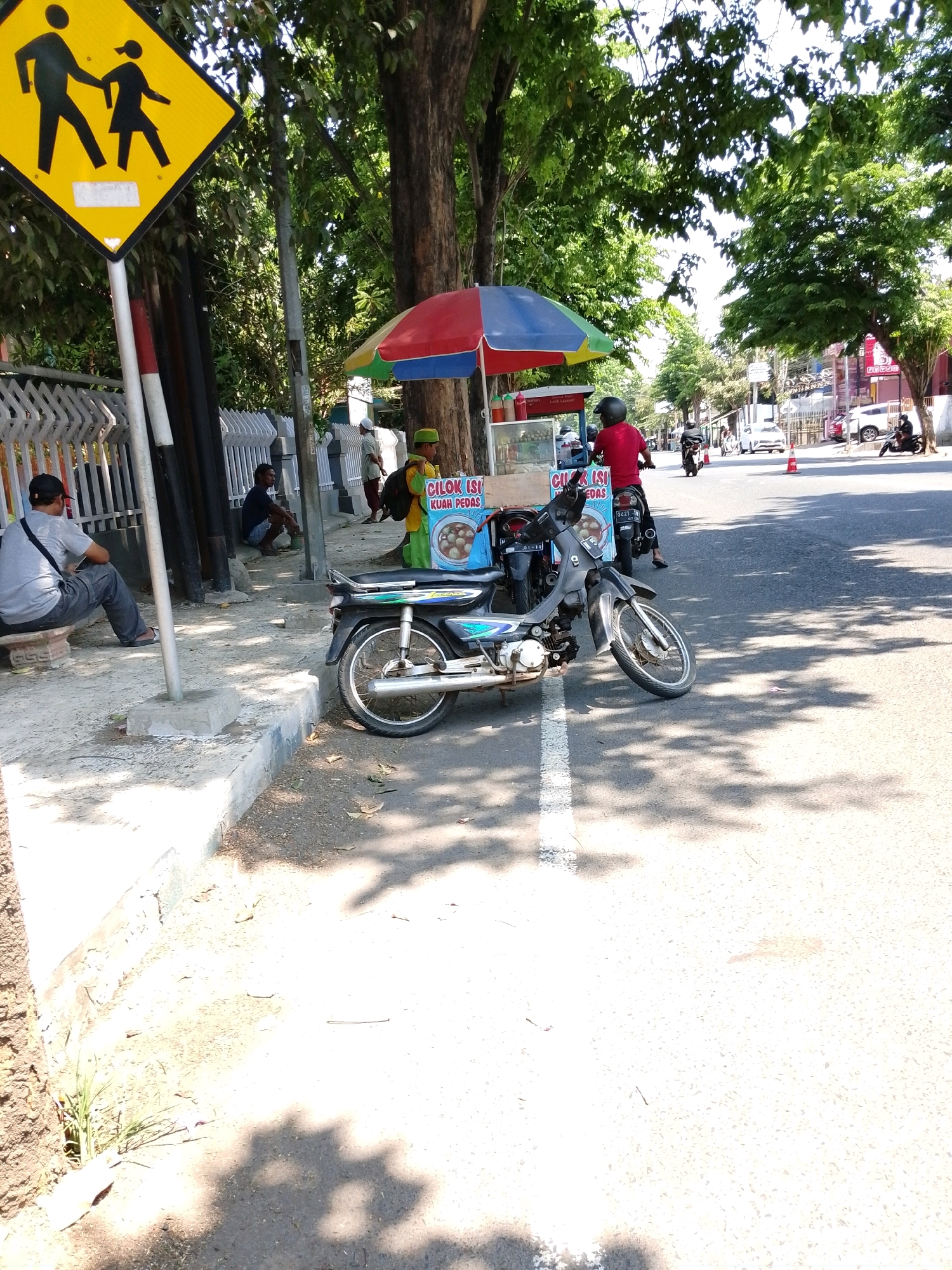 The image shows a street scene in Pasuruan, Indonesia, under a bright, sunny sky with dappled shadows cast by trees. On the left, a pedestrian crossing sign, a yellow diamond with black silhouettes of an adult and child, is mounted on a pole. Beside it, a man wearing a grey t-shirt and dark pants sits on a low concrete bench, looking towards the road. Further down the sidewalk, another person squats near a fence.
In the foreground, a parked motorcycle with a black frame and silver accents is angled towards the street. Behind the motorcycle, a food cart is set up under a colorful umbrella. The cart has a display of food items and signage that appears to be in Indonesian. A person in a red shirt and helmet is seated on a motorcycle next to the cart, facing away from the camera.
The street itself is paved and has a white line marking. Traffic cones are visible further down the road, and several other motorcycles and vehicles are present in the distance, suggesting an active urban environment. Buildings line the right side of the street. The overall impression is a typical day in a bustling Indonesian city.
