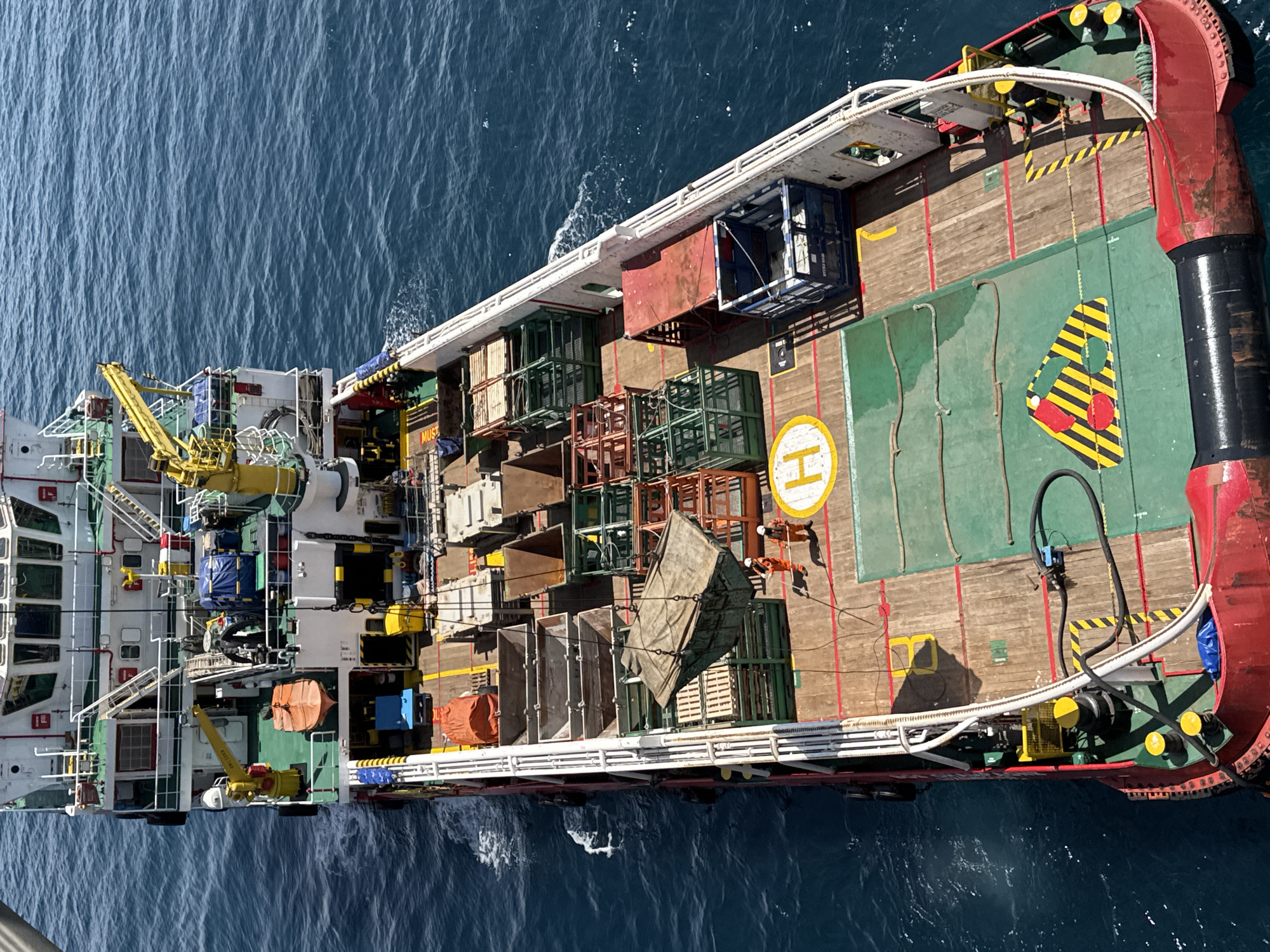 An aerial view captures the deck of a large vessel at sea. The vessel is red and white and appears to be a supply ship, with a helipad marked with a white "H" on the green deck. Various cargo, including metal cages, crates, and covered items, are visible on the deck. Two individuals in orange safety suits are standing near a large suspended load. The sea surrounding the vessel is a deep blue. There is no indication of the city of Unknown, Italy.