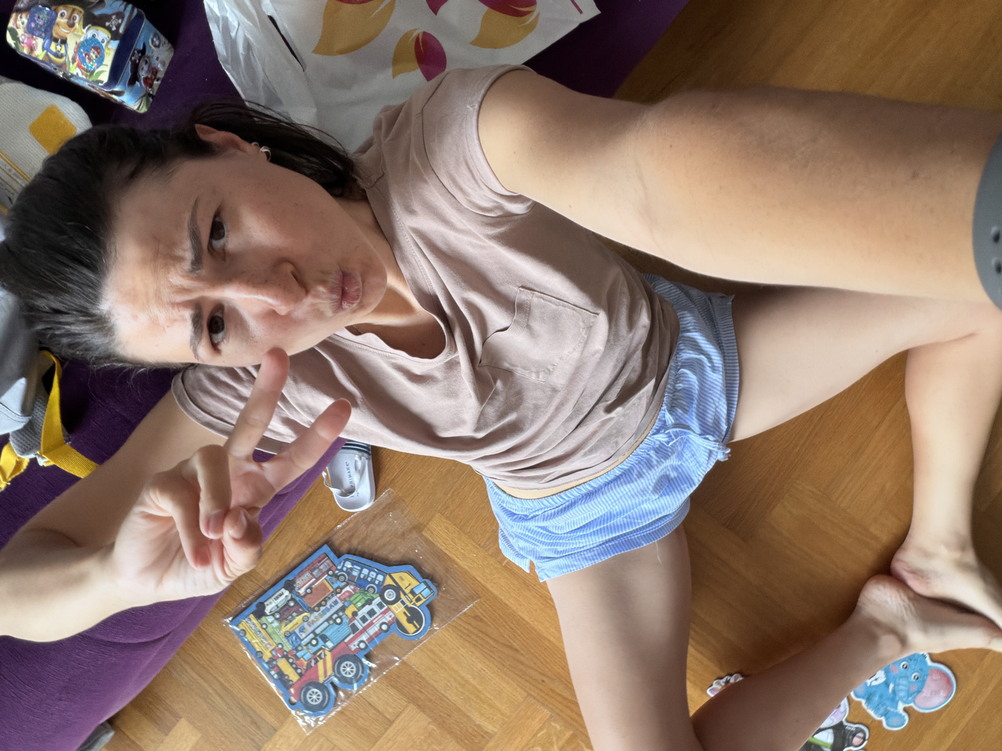 A woman lies on her back on a wooden floor. She wears a light pink t-shirt and blue and white striped shorts. She holds her hand up in a peace sign and makes a puckered face. A puzzle featuring various vehicles sits to her left. A white container labeled "PRIMARK" is adjacent to the puzzle. Her location in Budva, Montenegro, cannot be determined.