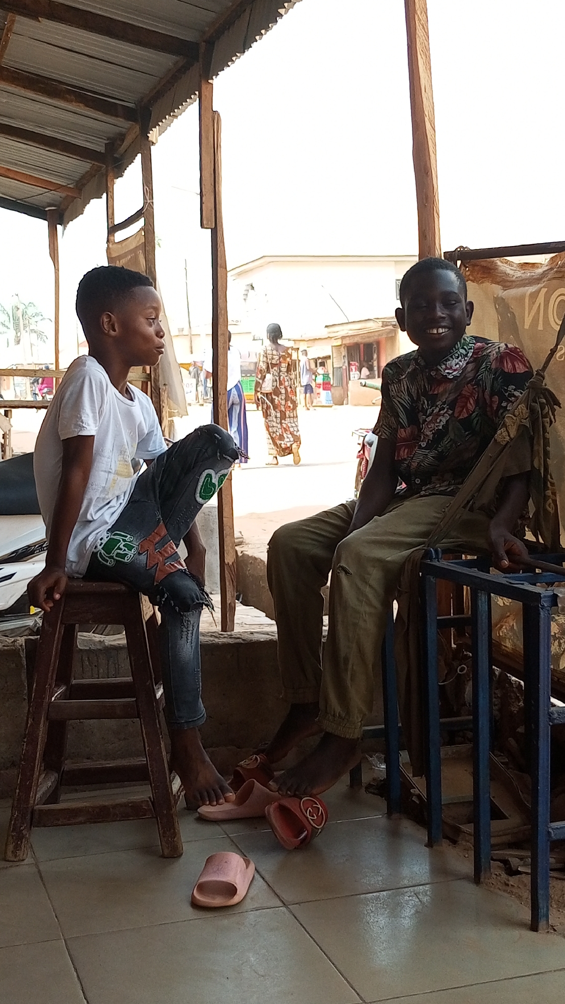 Two young males are sitting outside under a tin roof. The male on the left is sitting on a wooden stool with his legs crossed, he is wearing a white t-shirt and ripped jeans. The male on the right is wearing a colorful, leafy shirt and khaki pants. Three sandals are on the floor between them. Beyond the roof and poles, an unknown city street is visible with people walking along the street.